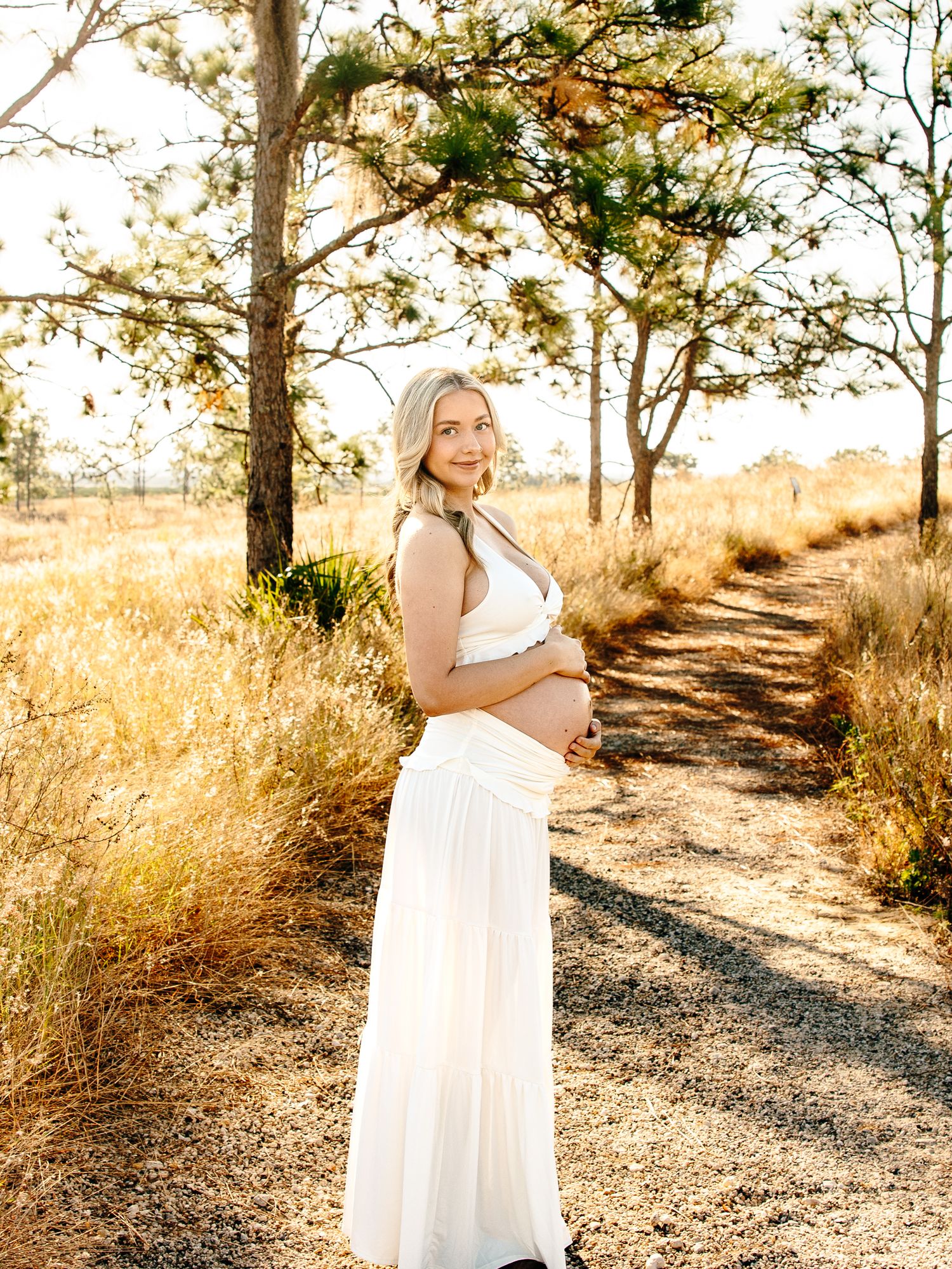 Pregnant woman in white dress holding baby bump on a sunlit dirt path surrounded by golden grass and trees.