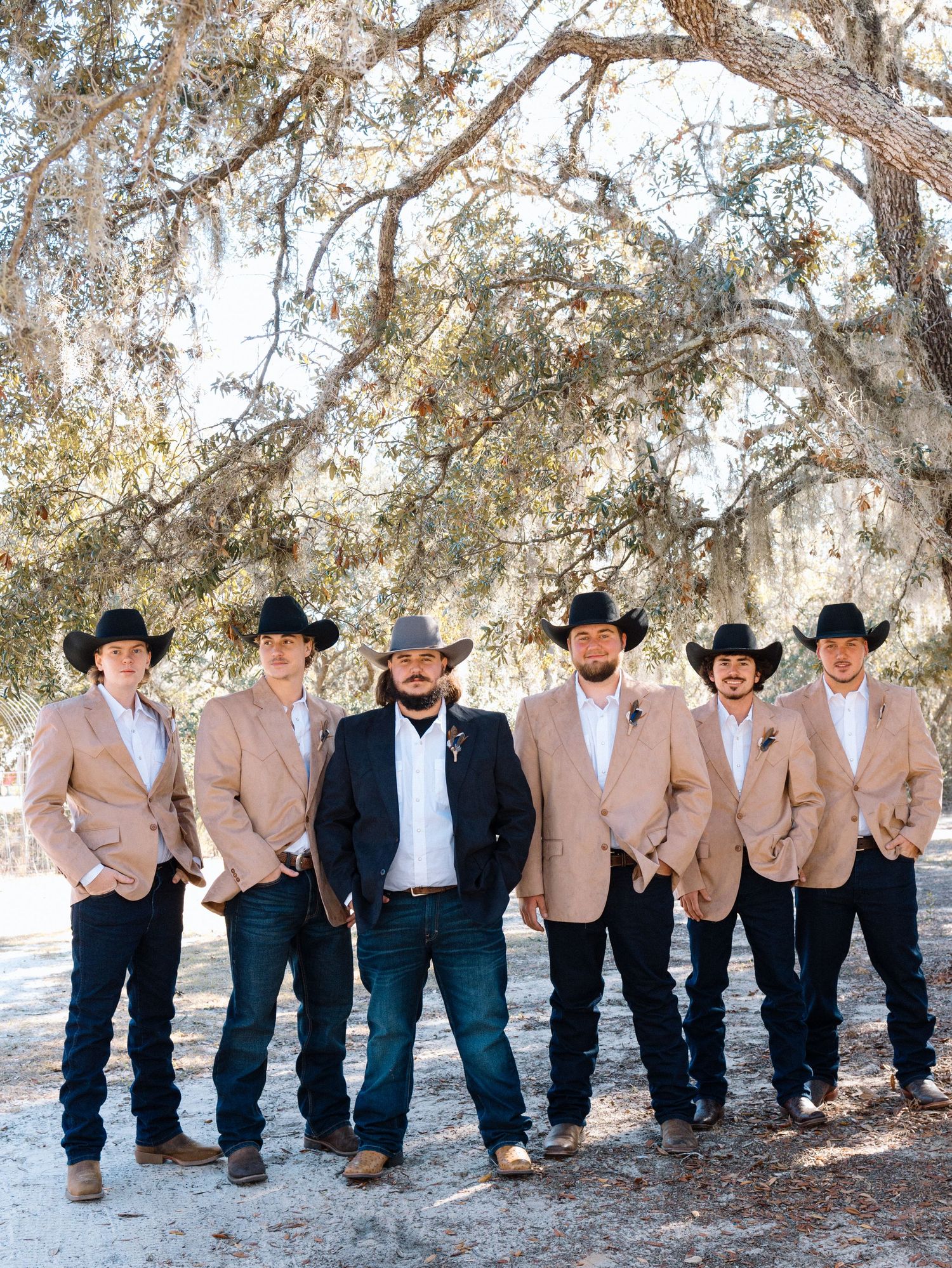 Group of groomsmen in matching beige jackets and black cowboy hats leaning against tree in casual outdoor setting.