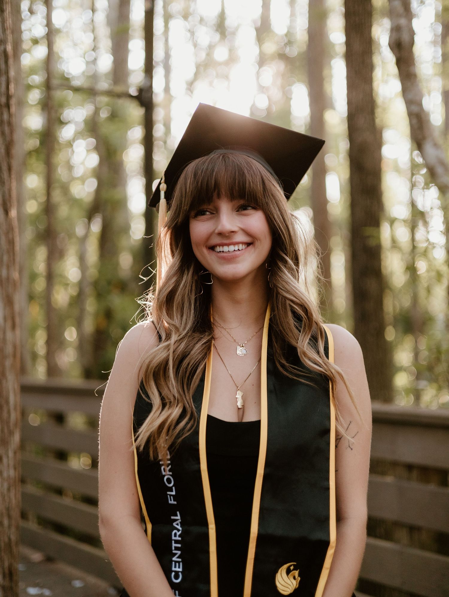 Graduate in cap and gown smiling in sunlit forest setting.