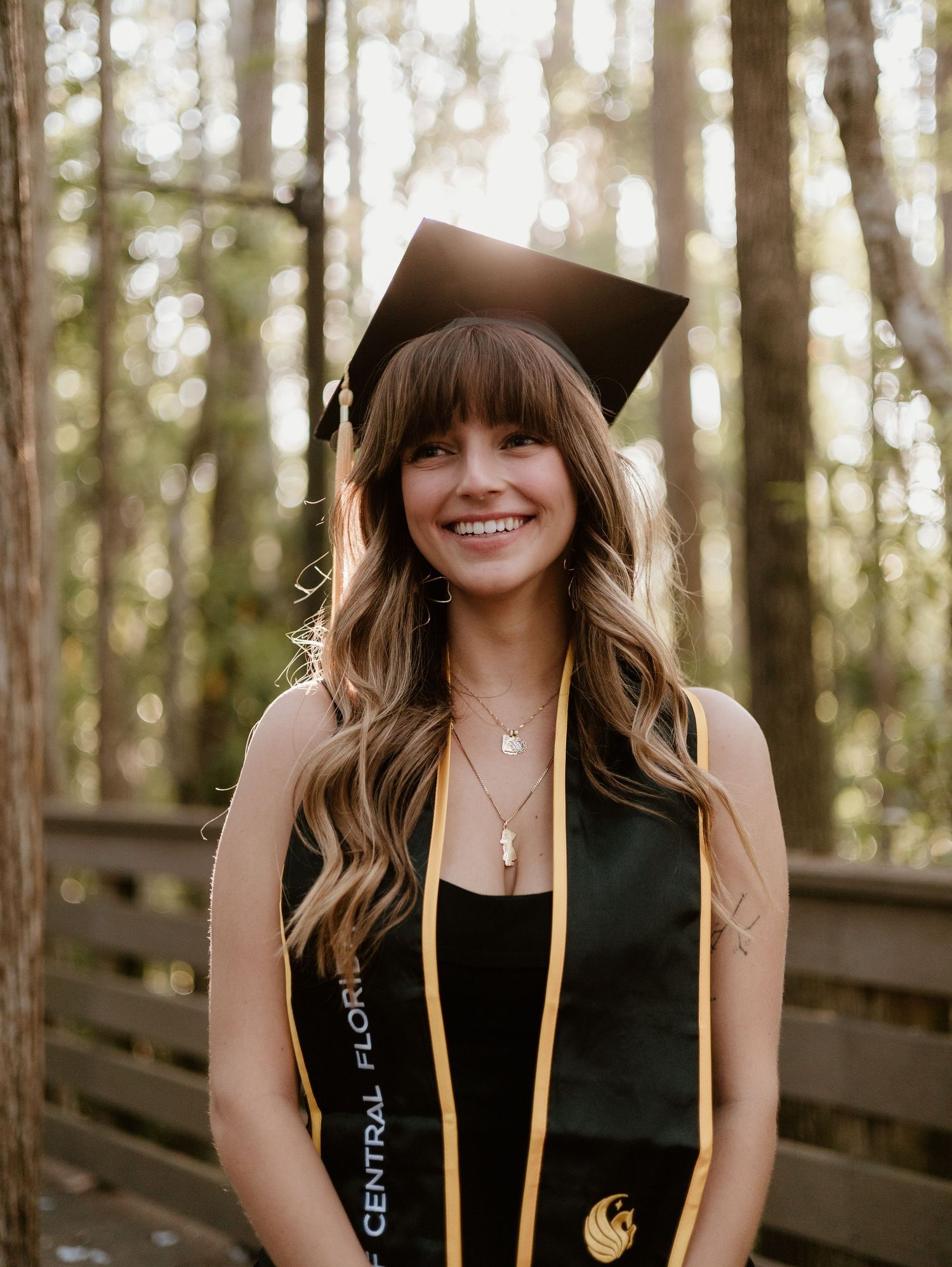 Smiling female graduate wearing black cap and UCF stole poses outdoors among trees on wooden bridge.