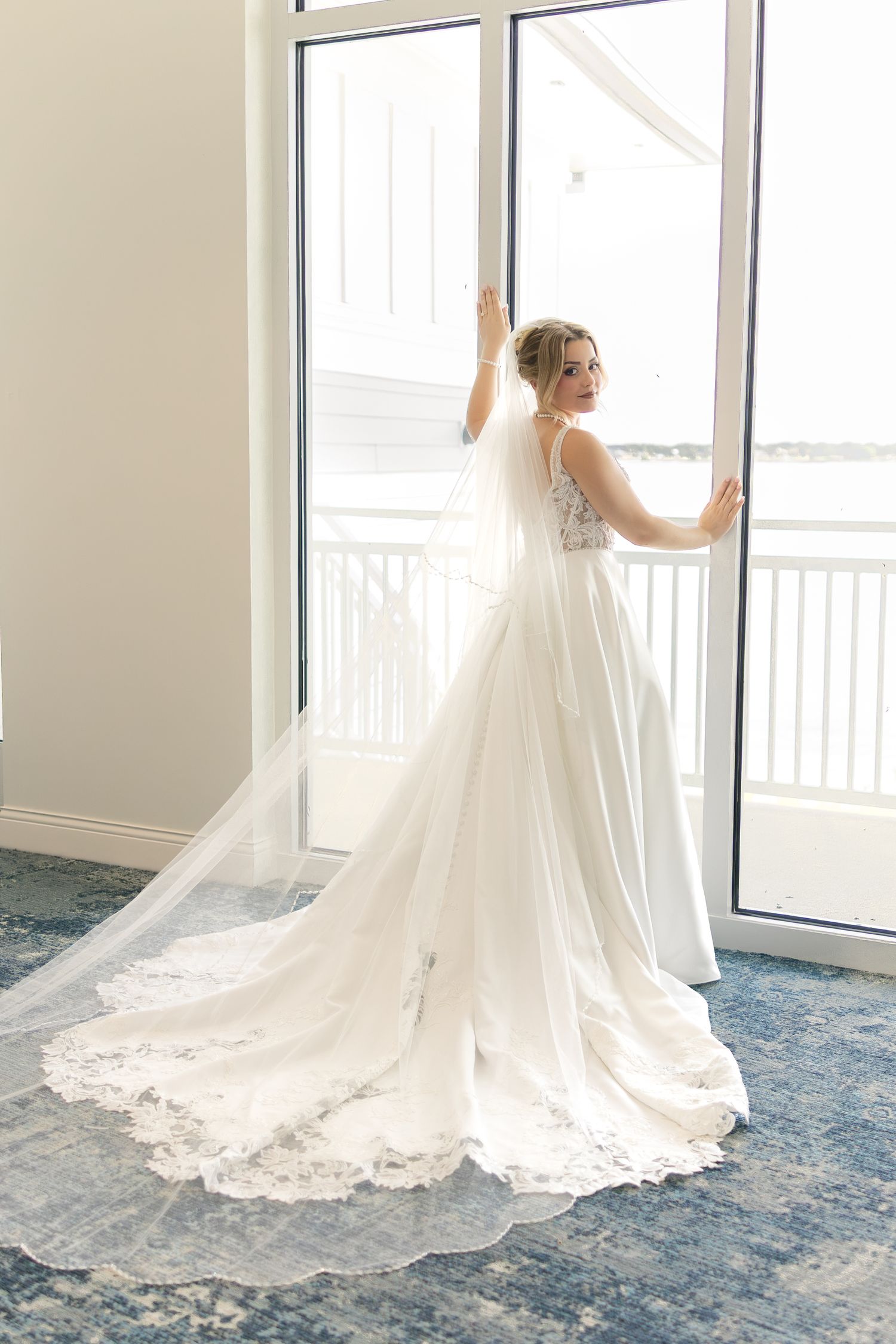 Bride in elegant white lace wedding gown with long veil and train stands by large window in bright room.