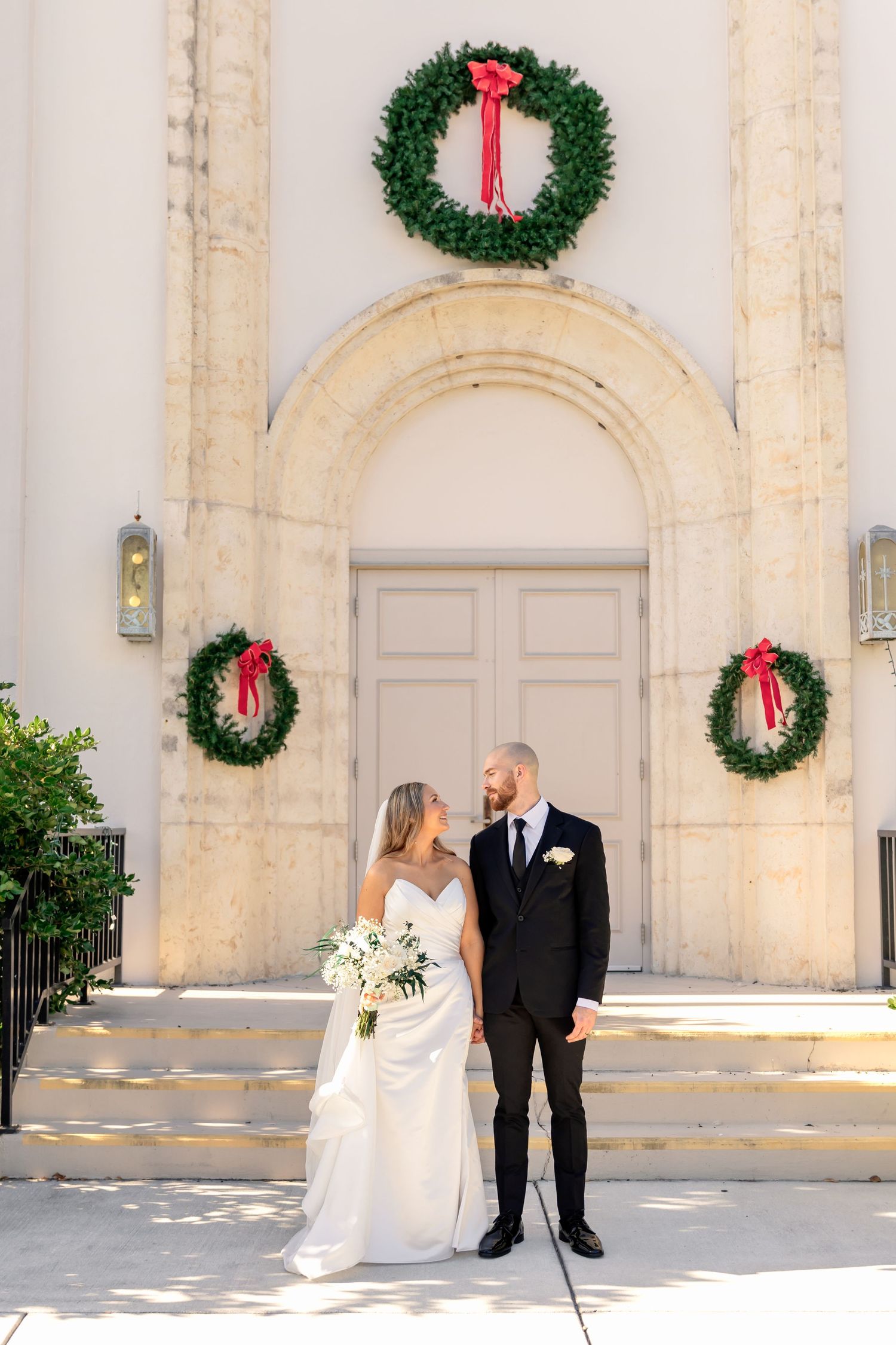 Bride and groom pose before a church entrance adorned with Christmas wreaths and red ribbons on their wedding day.
