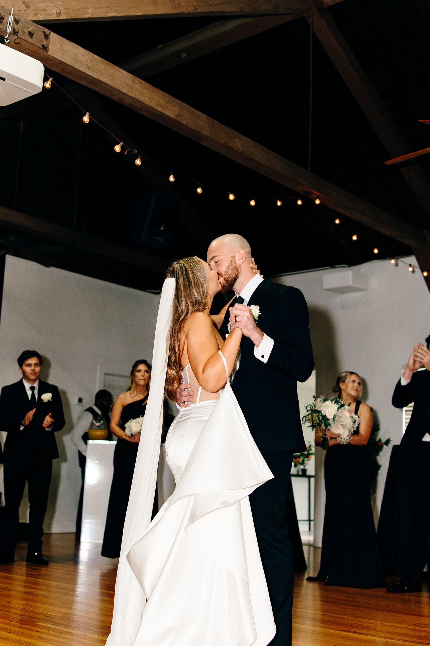 Bride and groom share first dance kiss in elegant venue with string lights and wedding party applauding behind them.