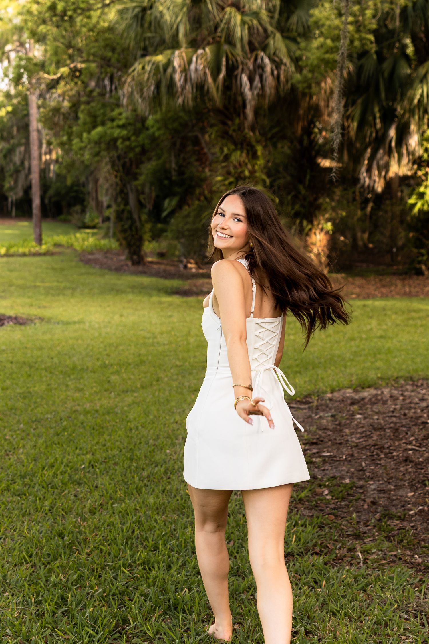 Young woman in white lace-back mini dress smiling over shoulder on green lawn with tropical trees.