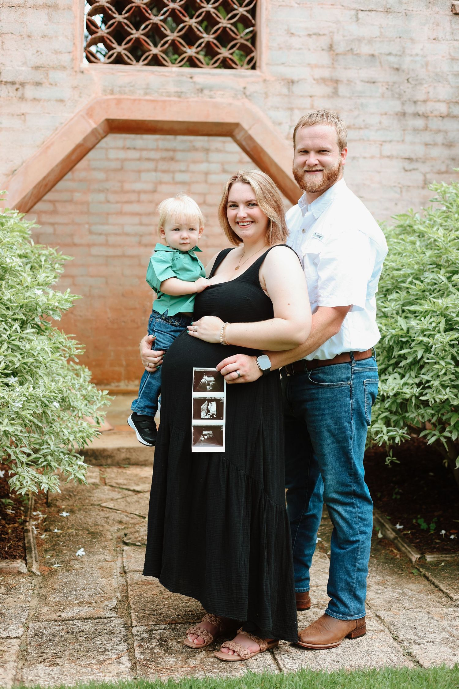 Family maternity photo with toddler in front of brick archway displaying ultrasound photos.
