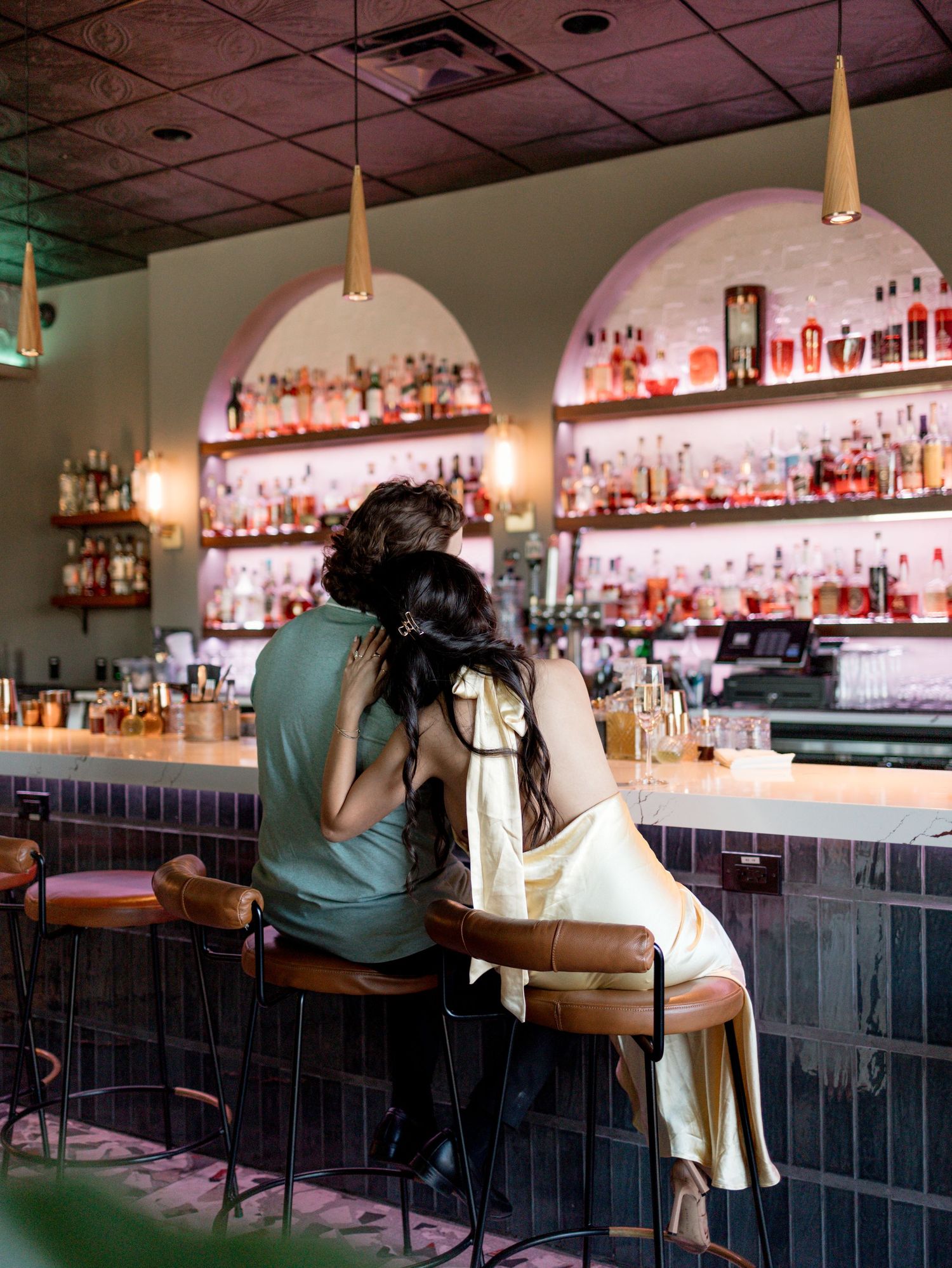 A cozy bar interior with illuminated arched shelving displays rows of liquor bottles while patrons sit at the counter.