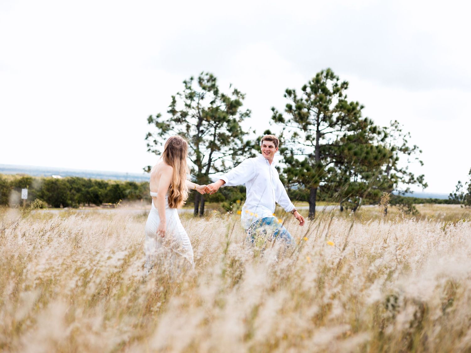 A couple in casual attire walks together through a sunlit field of tall grass on a summer day.