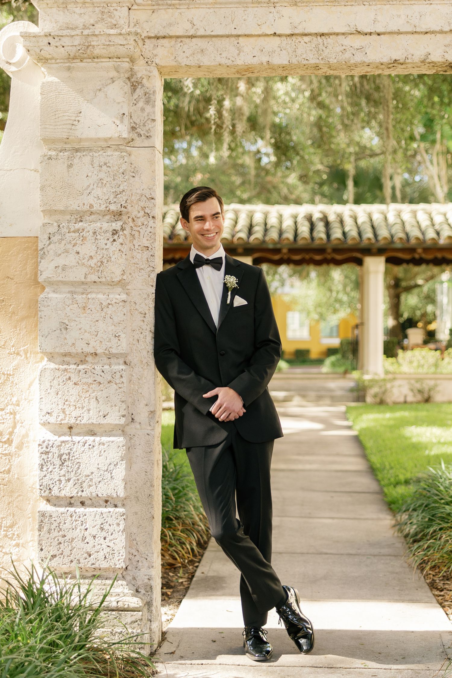 Smiling groom in black tuxedo with bow tie and boutonniere leaning against stone pillar at outdoor wedding venue.