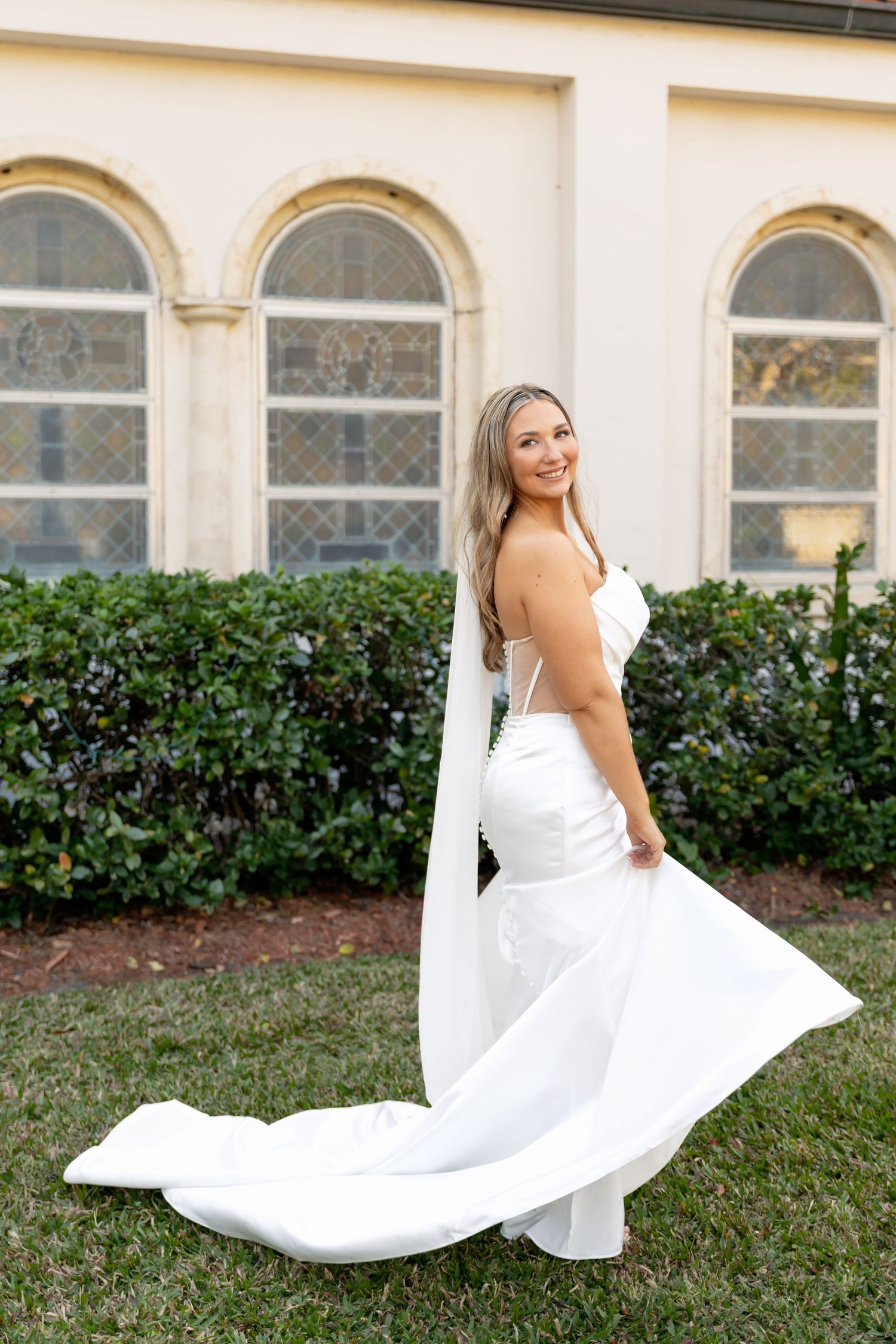 Bride in white mermaid gown with flowing train poses outdoors near elegant arched windows and lush hedges.