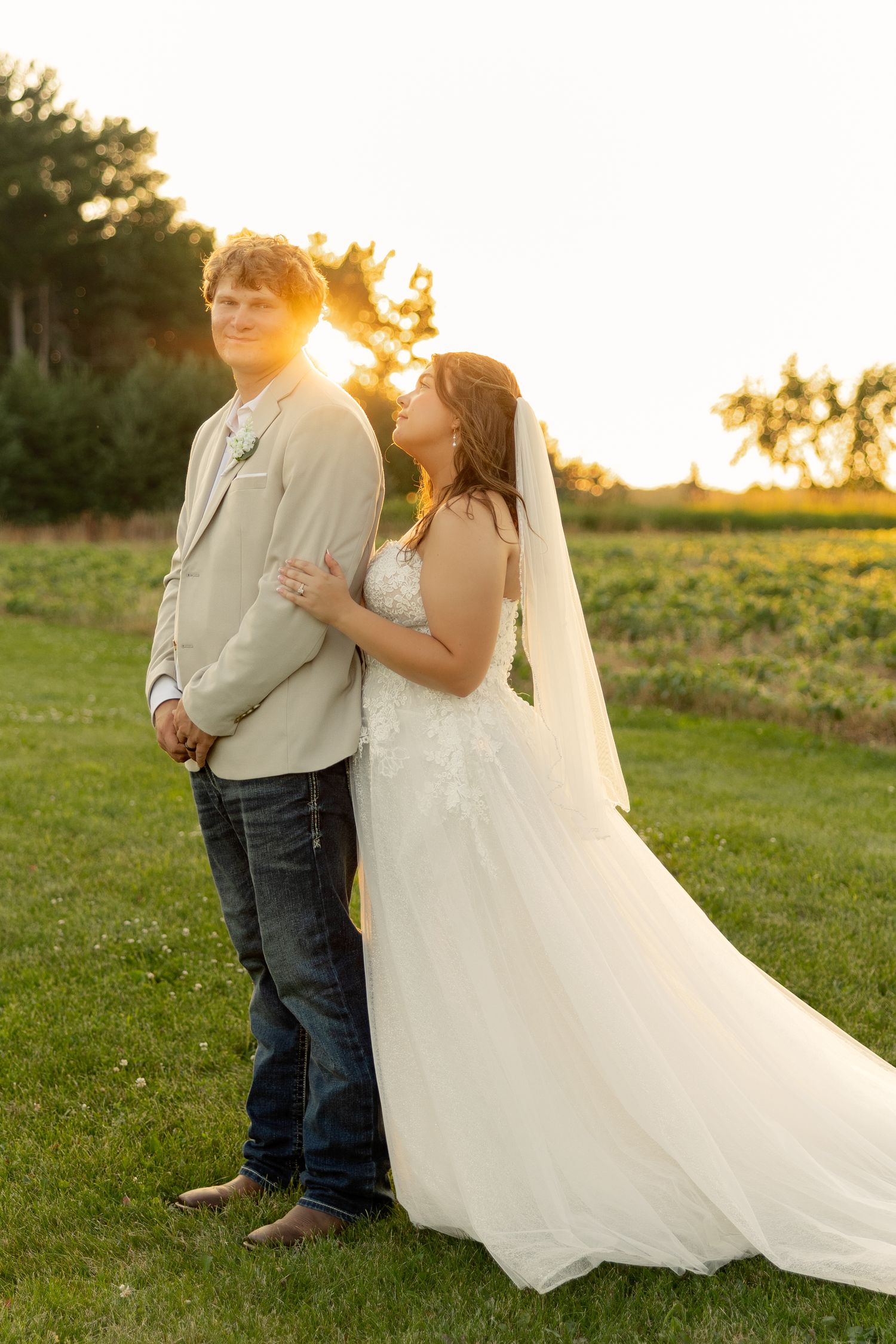Bride in white gown and veil stands with groom in tan jacket and jeans during golden sunset outdoors.