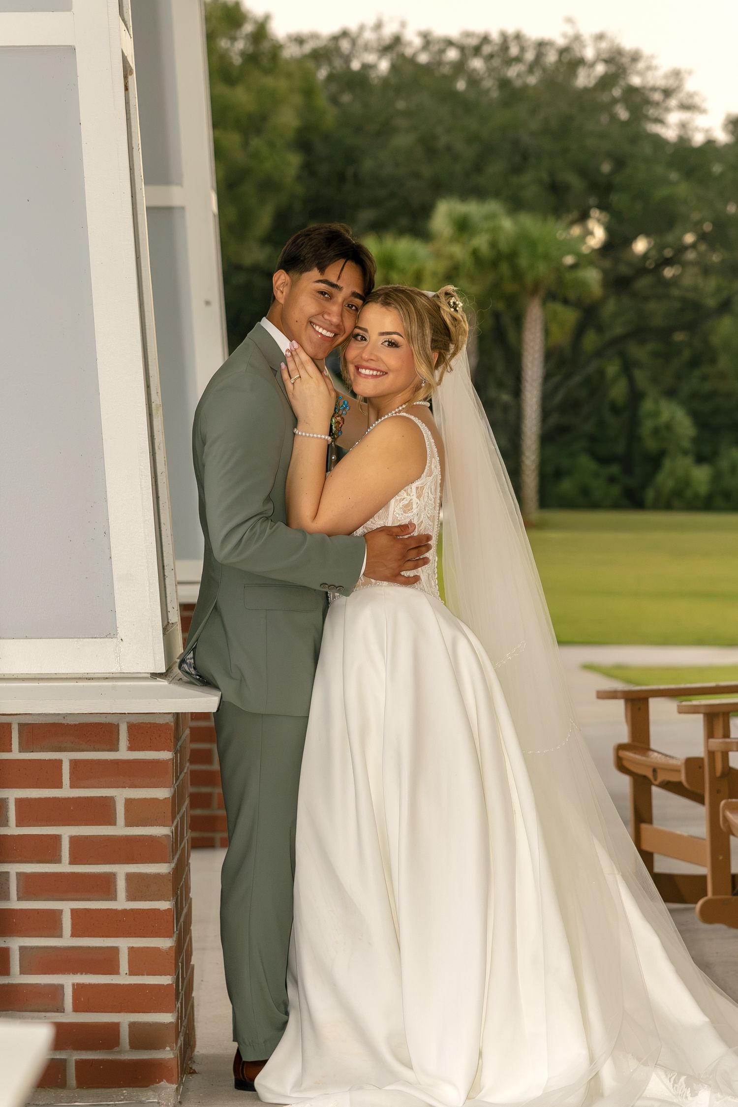 Bride in white gown and veil embraces groom in gray suit on a brick porch at an outdoor wedding venue.