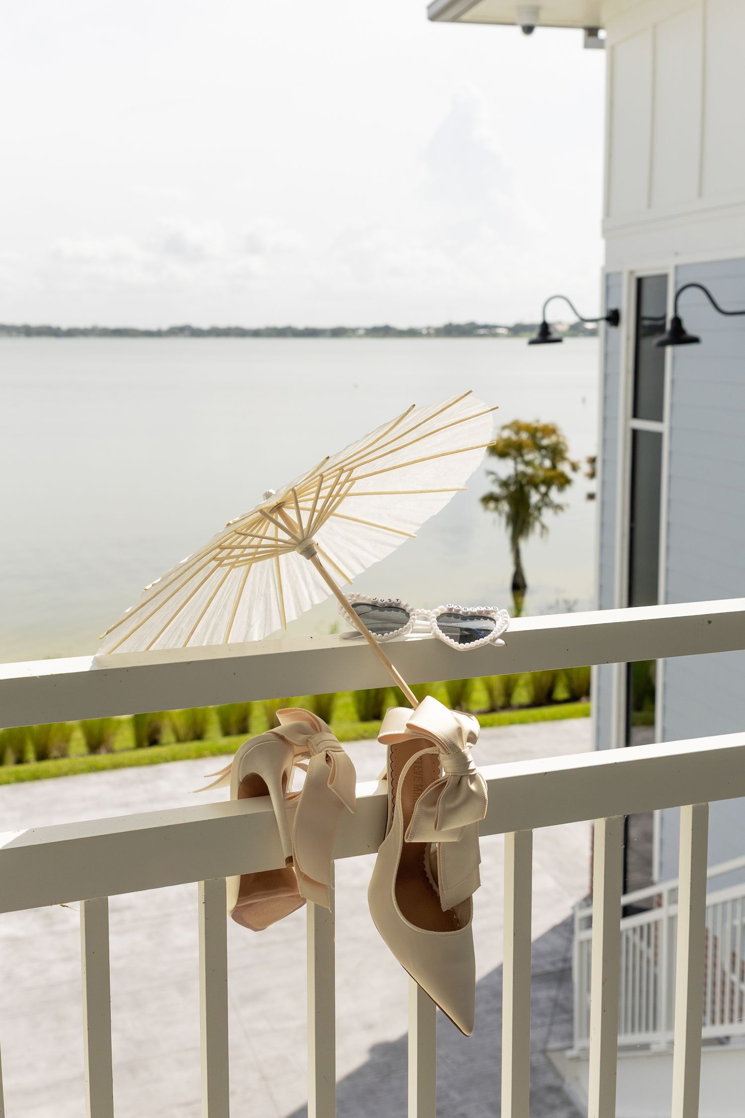 Elegant cream heels and sunglasses hang on a white balcony railing with a parasol overlooking a serene waterfront.
