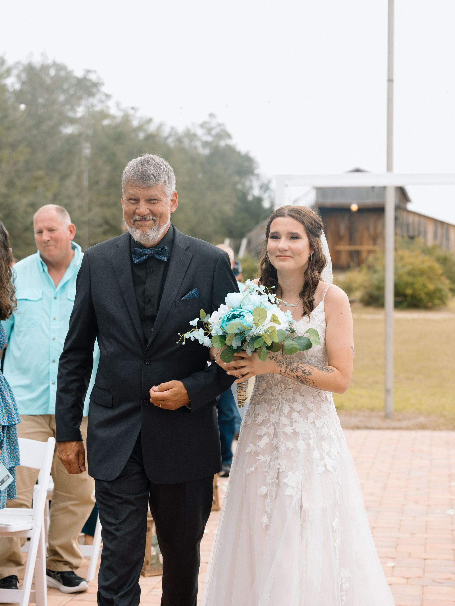 A bride in a white lace wedding dress walks down a brick path carrying a turquoise and white floral bouquet.