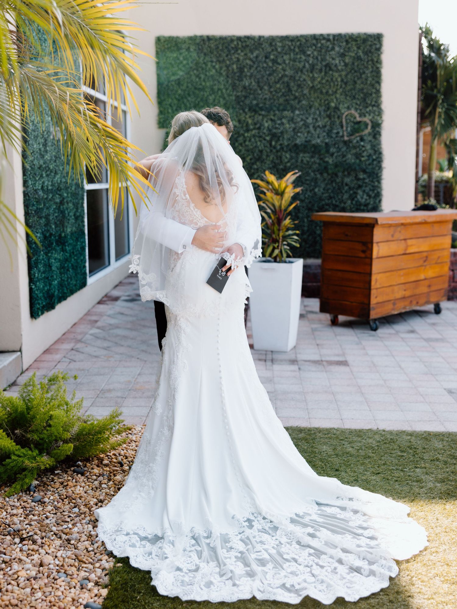 Bride in lace wedding gown with veil embracing groom in outdoor garden setting with greenery wall backdrop.