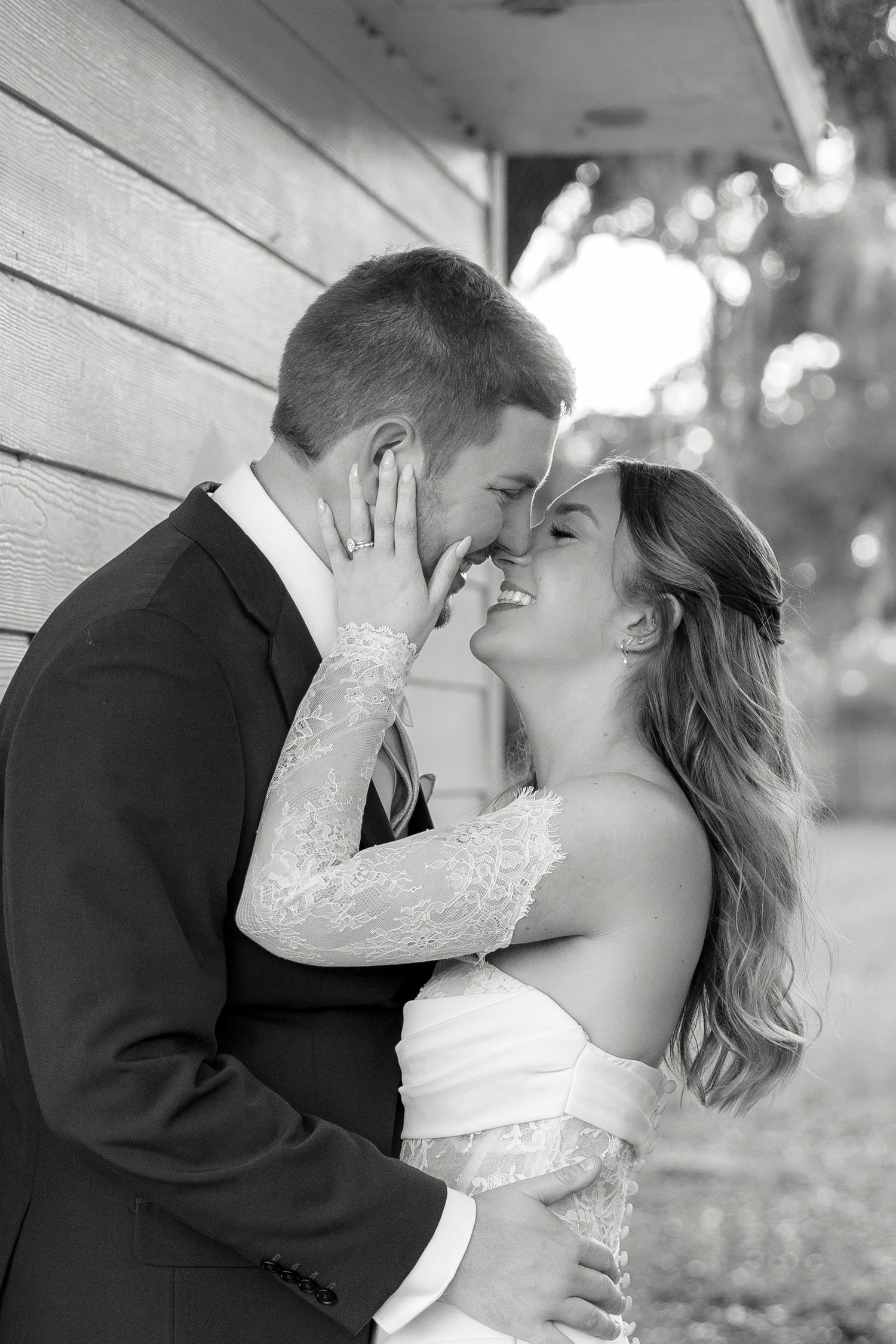 Black and white photo of a smiling bride and groom sharing a romantic almost-kiss on their wedding day.