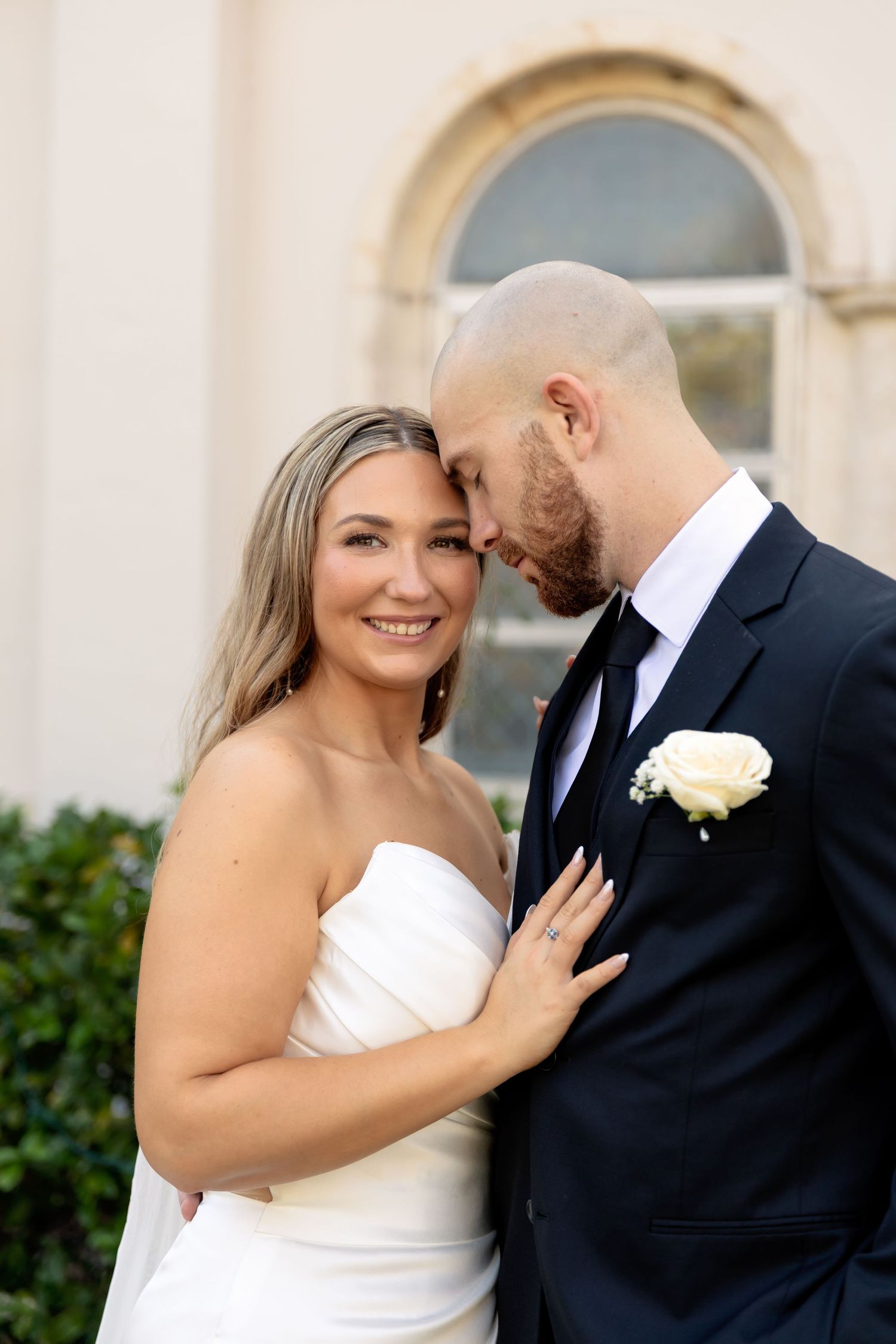 A smiling bride in a white strapless gown leans close to her groom in a dark suit with a white rose boutonniere.