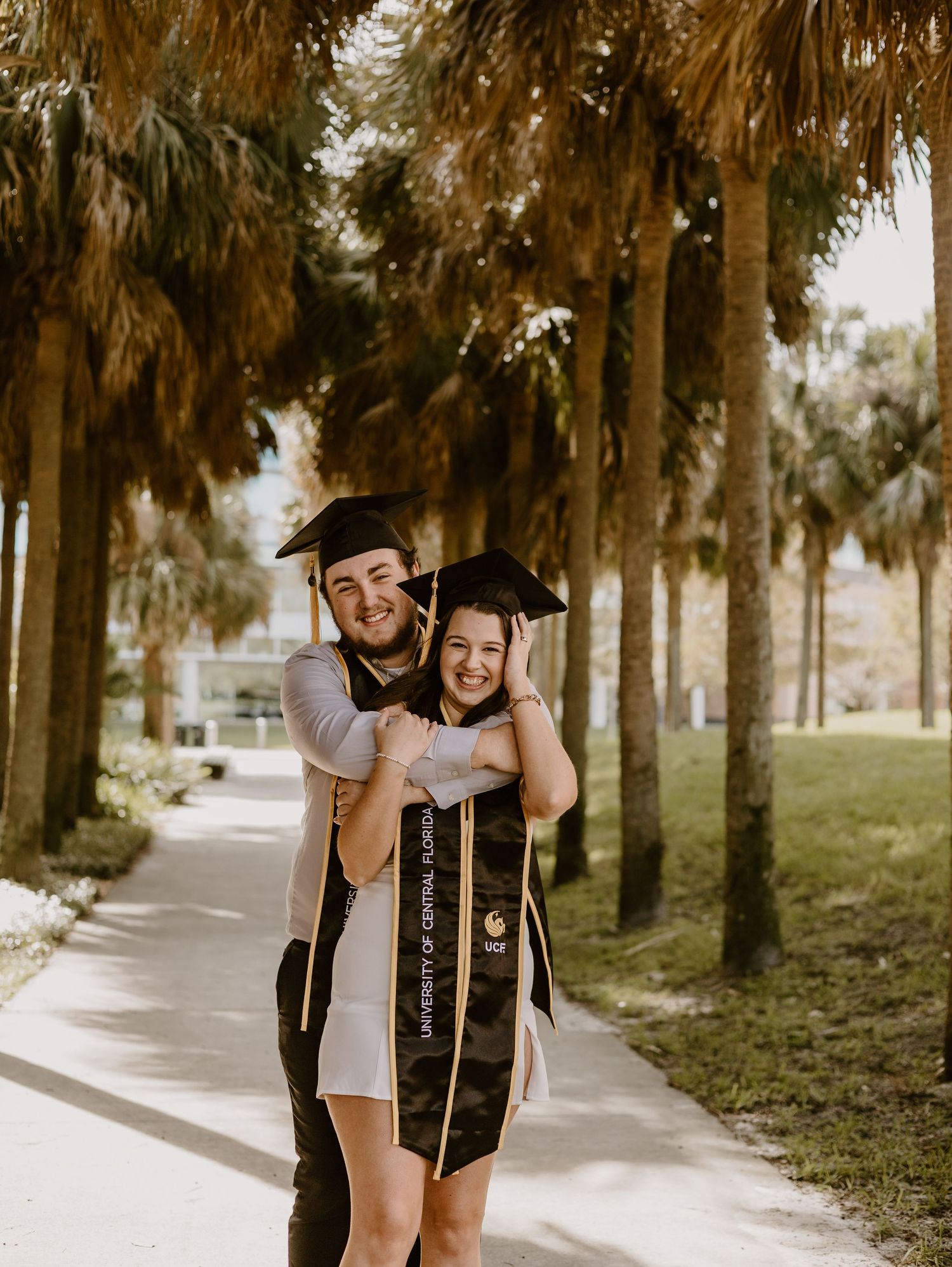 Two UCF graduates in caps and stoles embrace and smile on a palm tree-lined campus walkway.