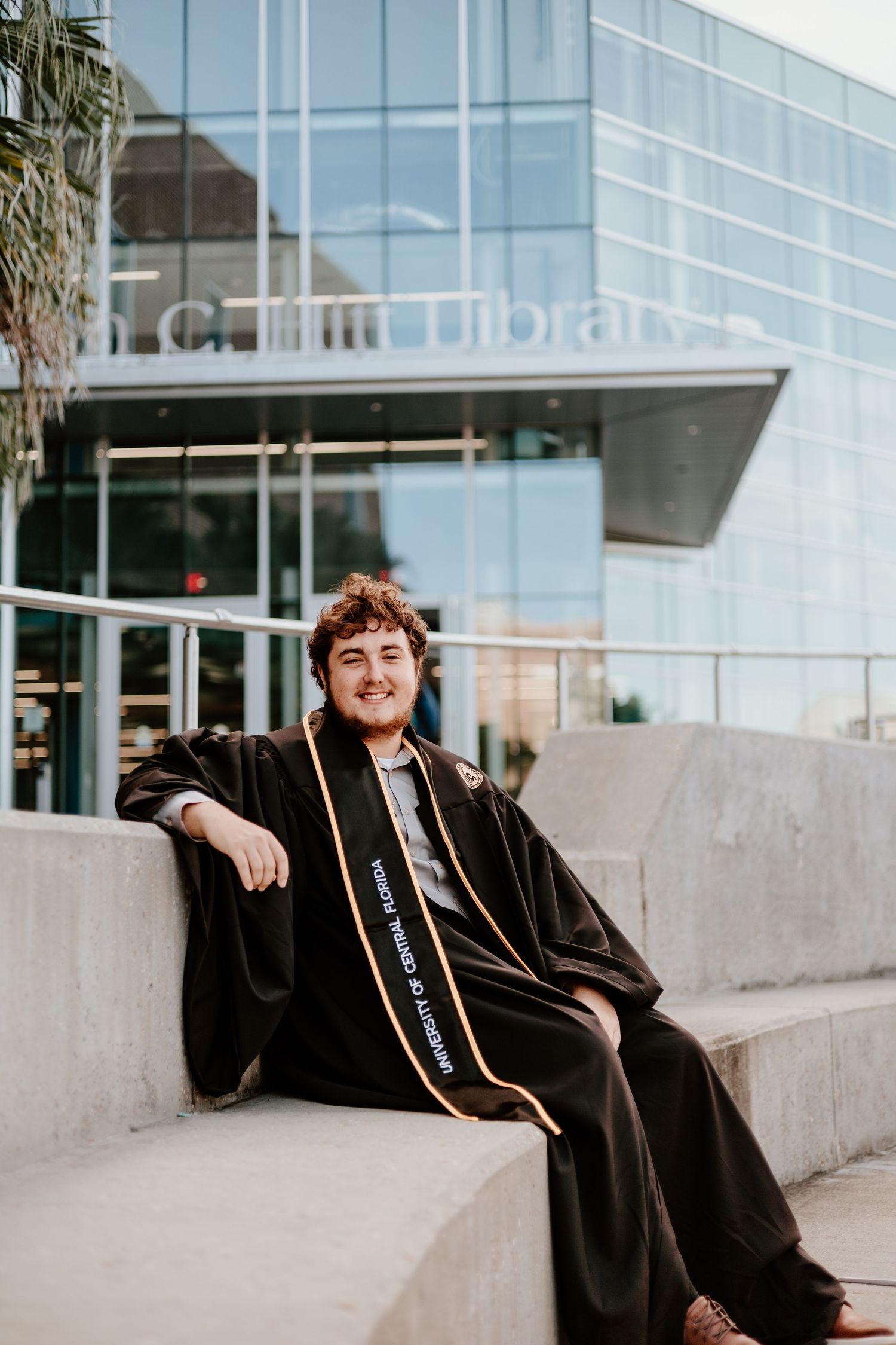 A smiling graduate in black cap and gown with gold sash sits on concrete steps outside a university library.