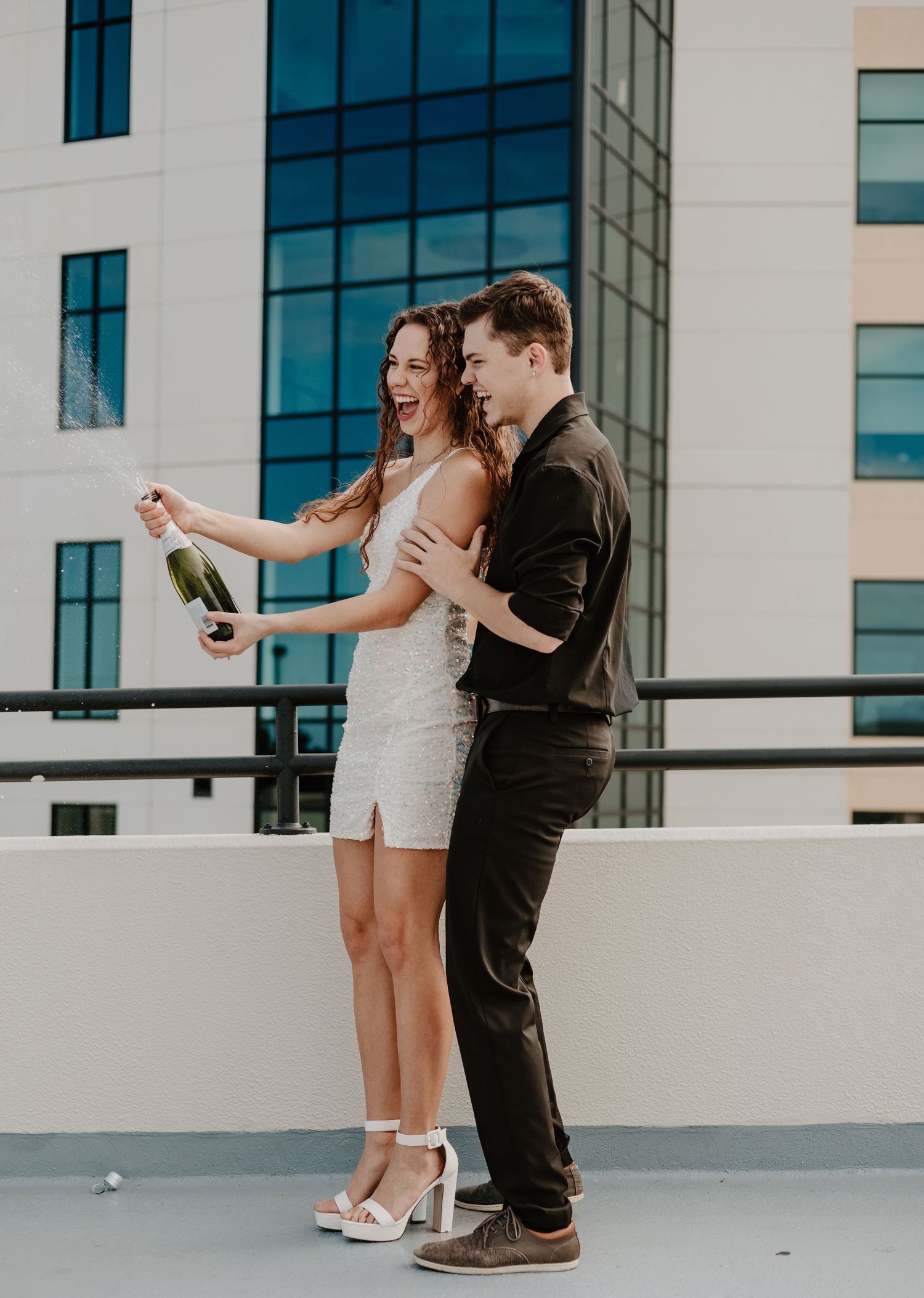 A couple opens champagne on a modern rooftop terrace with glass buildings in the background.