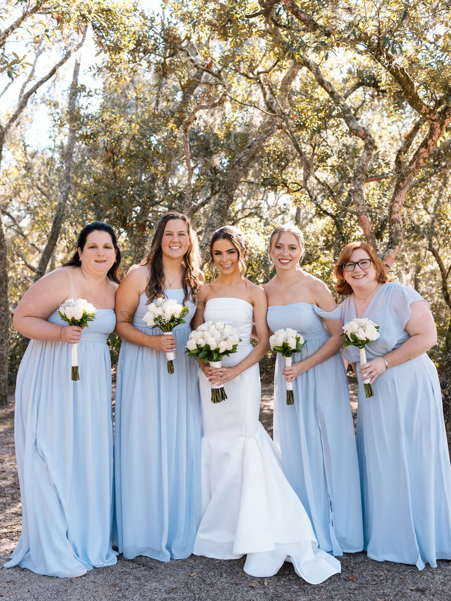 Wedding party wearing light blue bridesmaid dresses poses outdoors under sun-dappled trees in coordinated style.