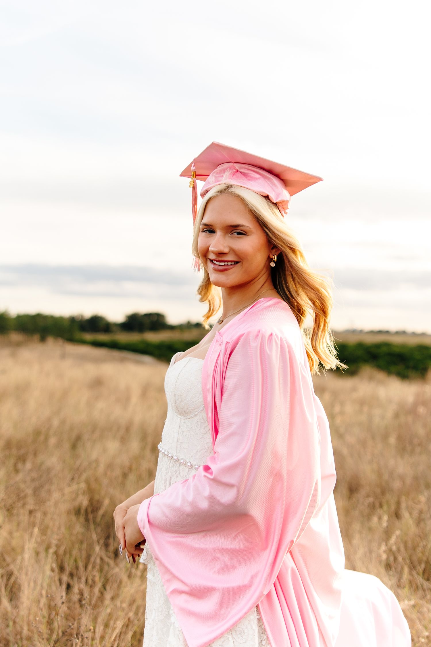 Smiling blonde graduate in pink cap and gown posing in a golden field under a cloudy sky.