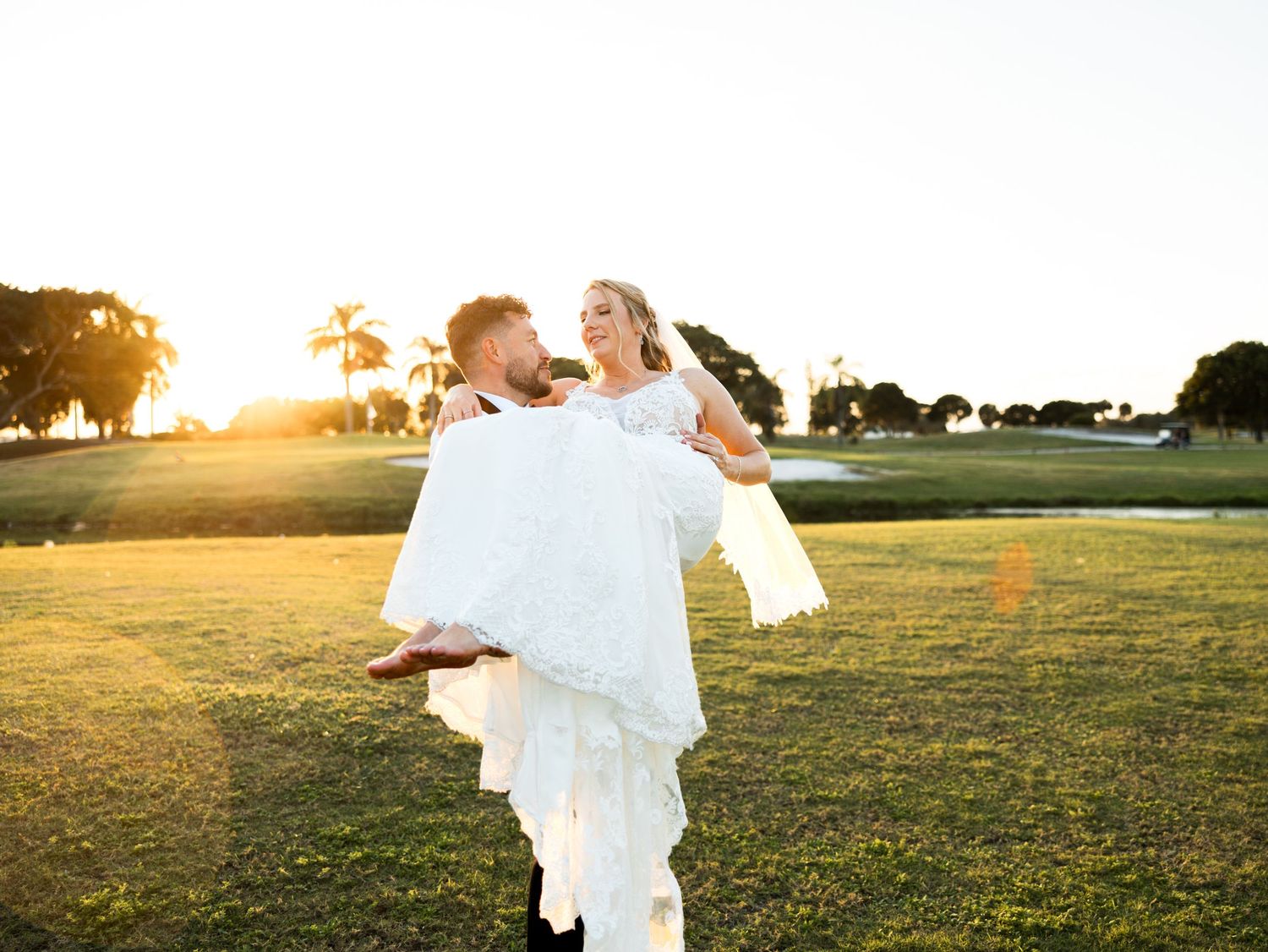Groom lifting bride in wedding dress at golden hour on a lush green golf course.