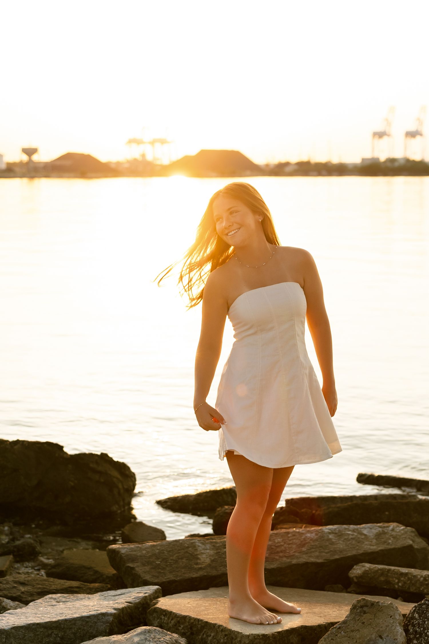 Smiling woman in white strapless dress standing on rocks by the water at golden sunset.