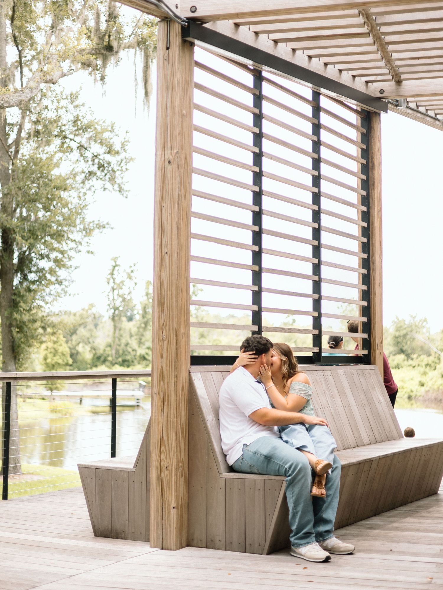 Couple sharing a romantic moment on a modern wooden bench under a pergola overlooking a serene waterfront.