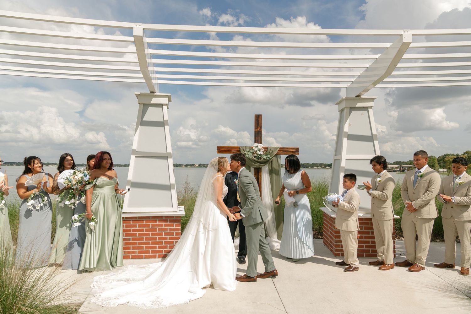 Outdoor wedding ceremony with bride and groom at altar surrounded by bridal party under pergola.