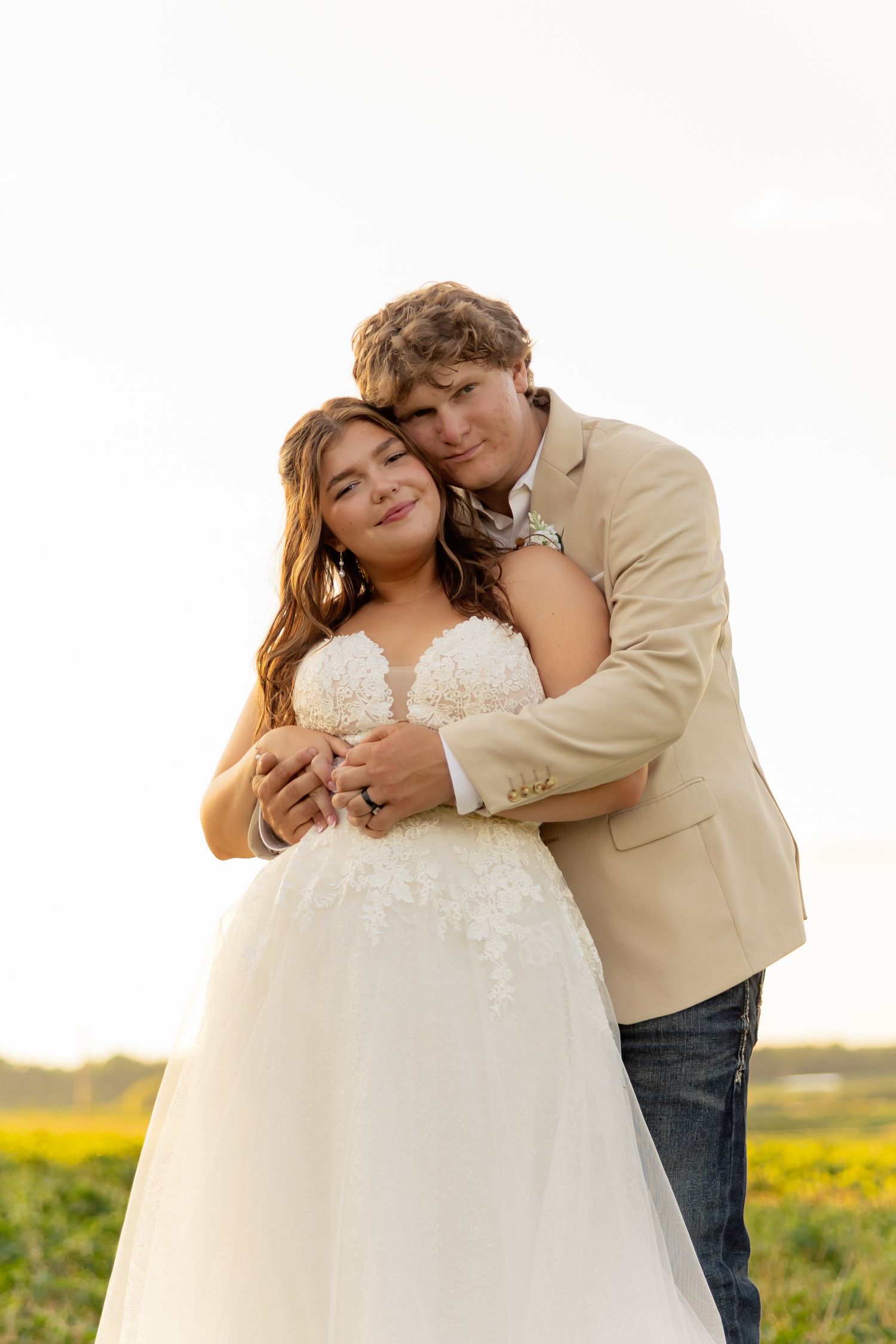 A couple in wedding attire embraces in a field, the bride in white lace gown and groom in tan blazer and jeans.