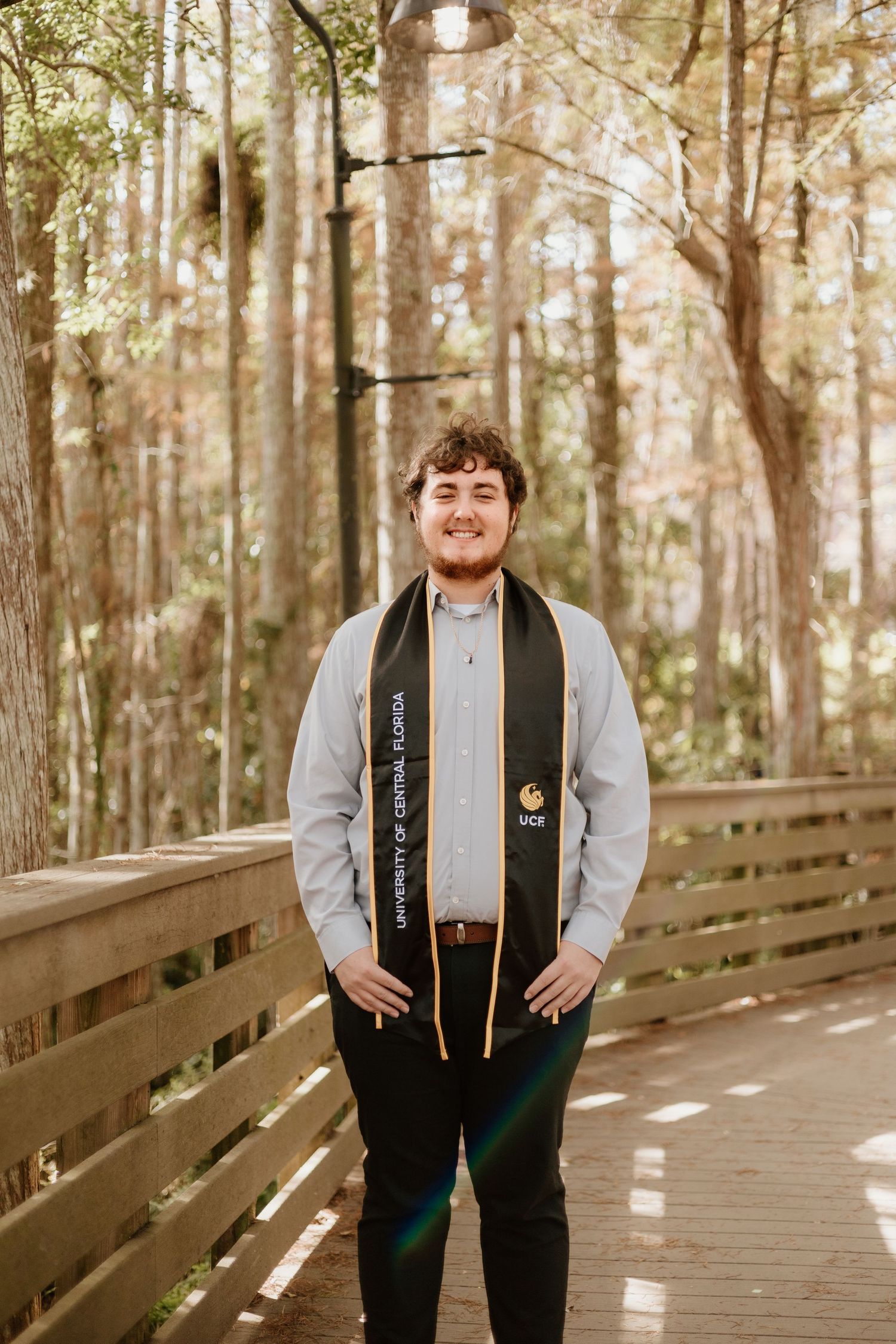 UCF graduate smiling on wooden boardwalk in forest, wearing University of Central Florida graduation stole.
