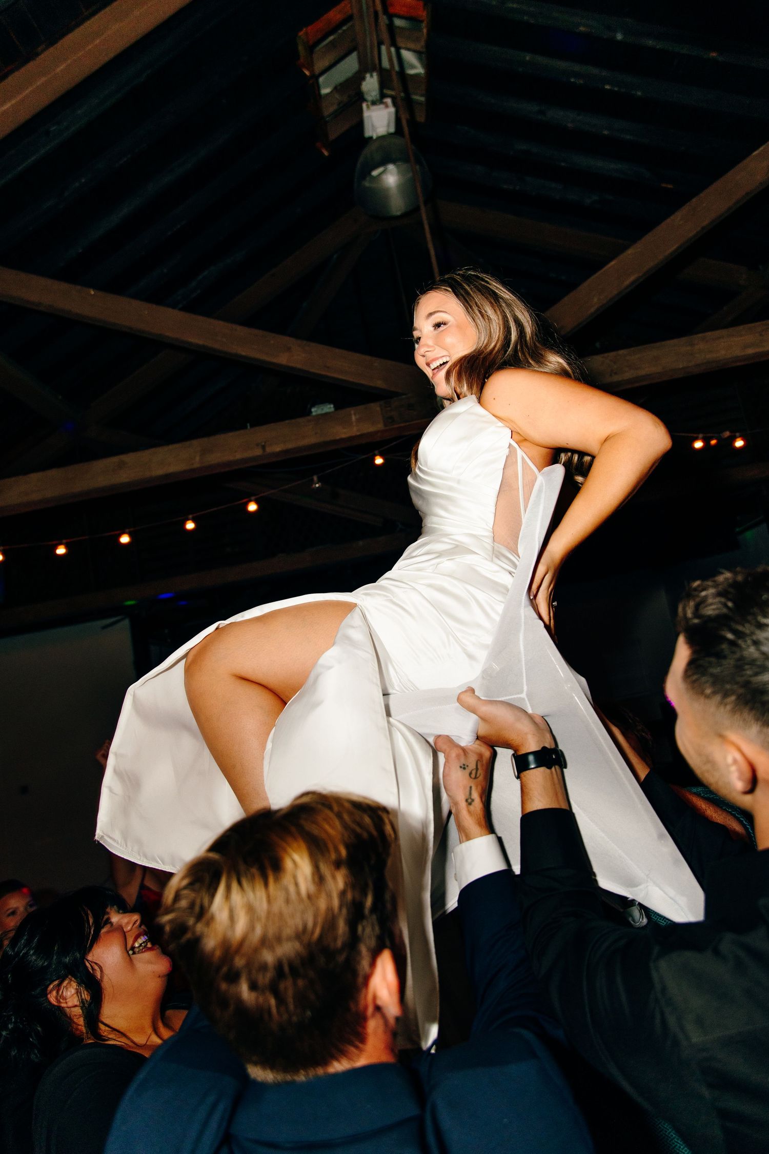 Bride in white dress lifted by guests during lively wedding reception celebration indoors.