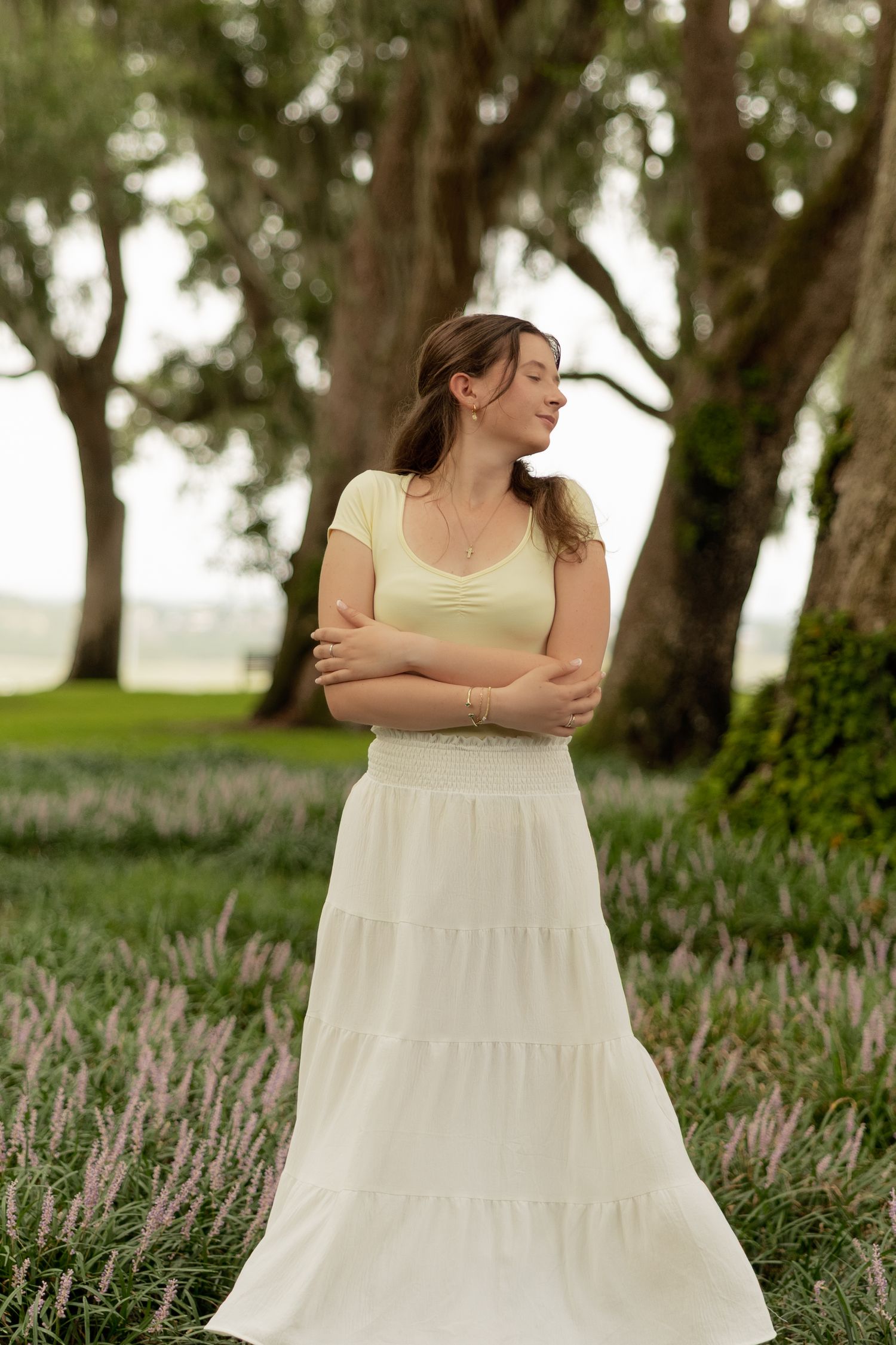 Woman in white tiered maxi skirt and yellow top standing in a flower field with trees in background.
