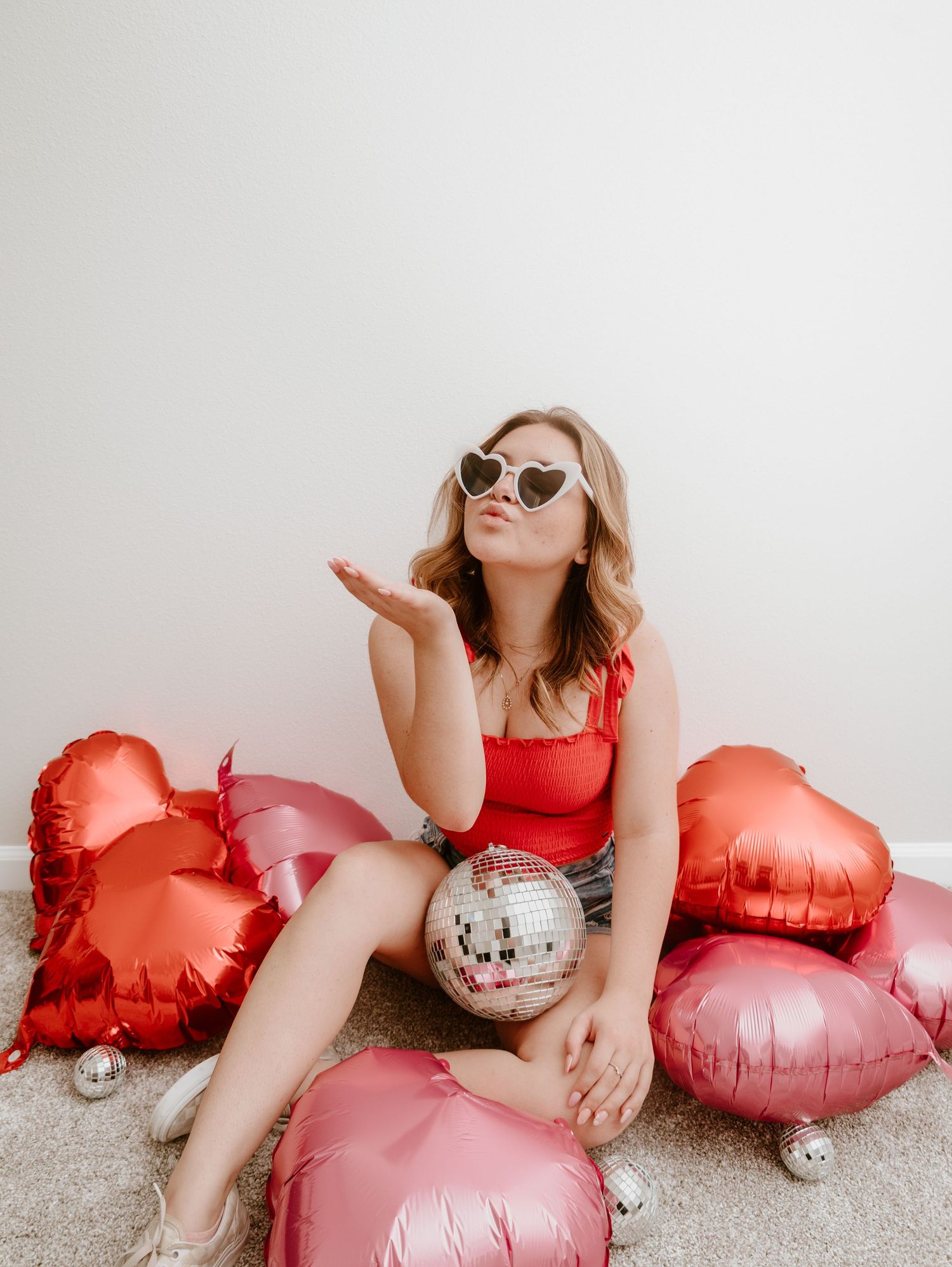 A person in red lounges on the floor with pink and red heart-shaped balloons and a disco ball in a Valentine's Day setting.