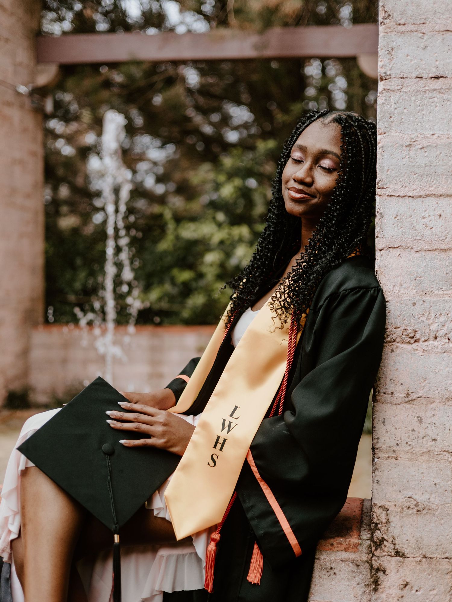 A graduate in academic regalia sits on stone steps while holding their diploma during a graduation photoshoot.