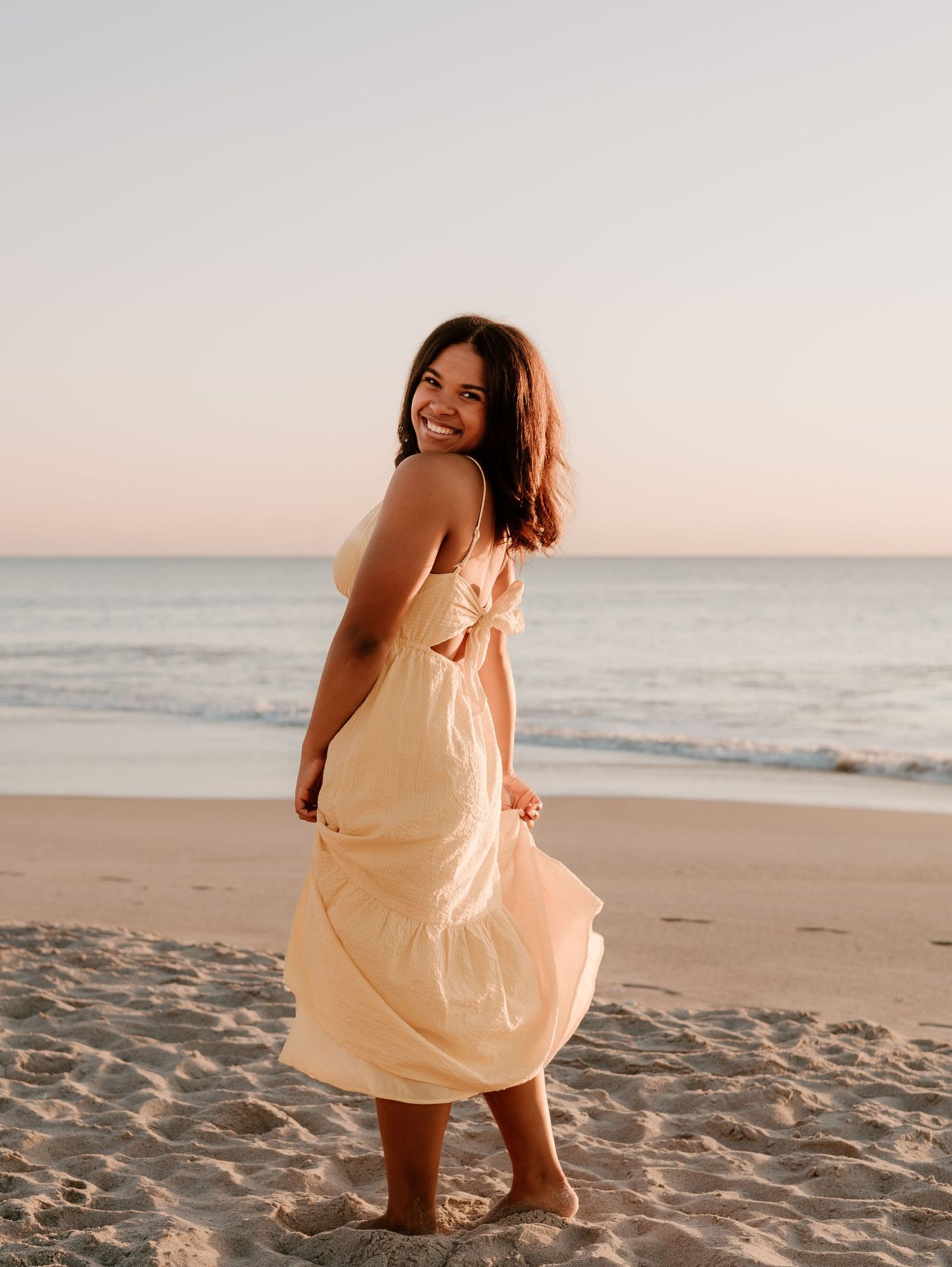 A person in a peach-colored dress twirls joyfully on a sandy beach at sunset with ocean waves in the background.