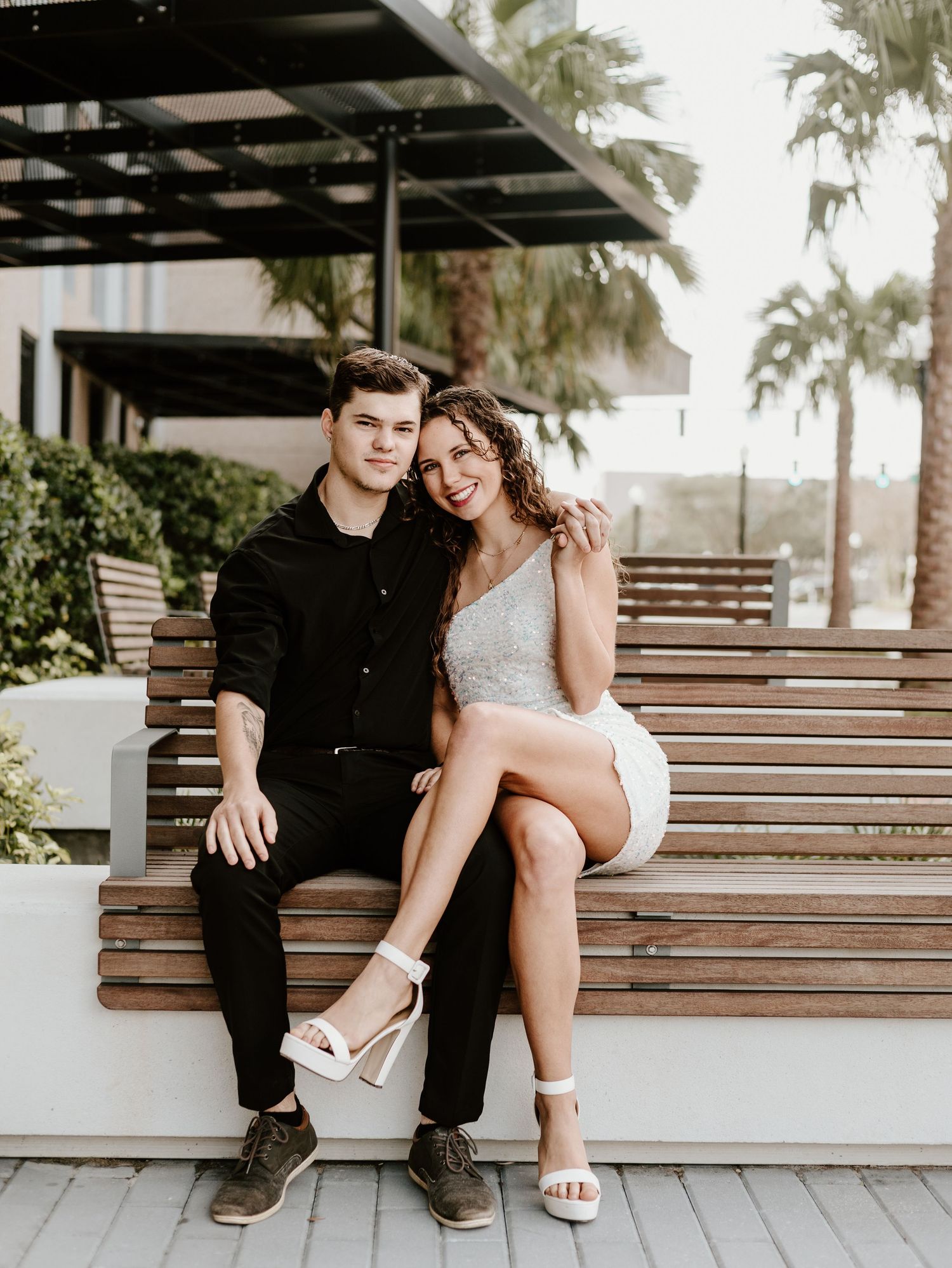 A couple sits casually together on wooden steps outdoors with palm trees visible in the background.