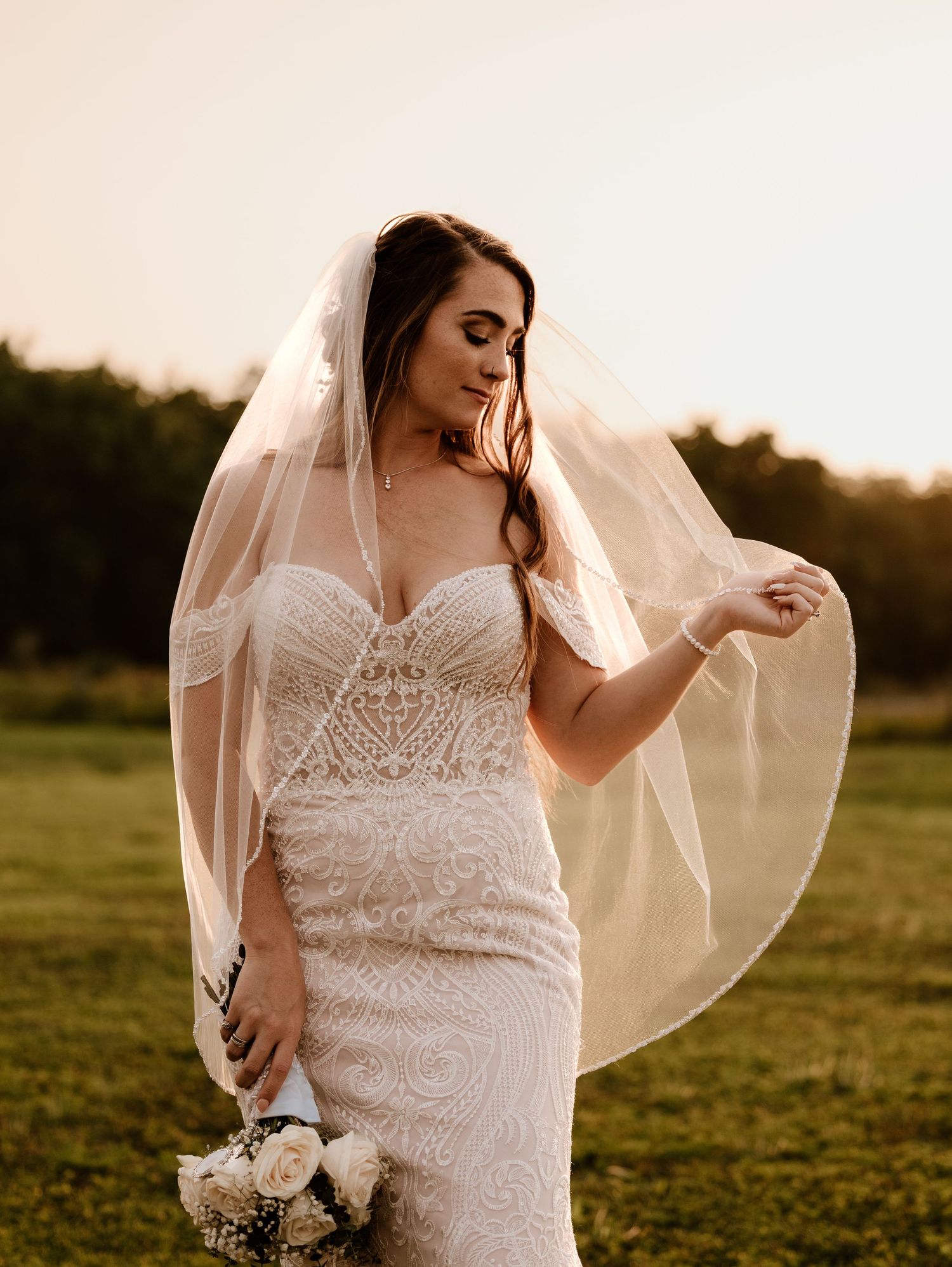 A bride in a lace gown poses in a golden sunlit field during sunset with her veil flowing in the breeze.