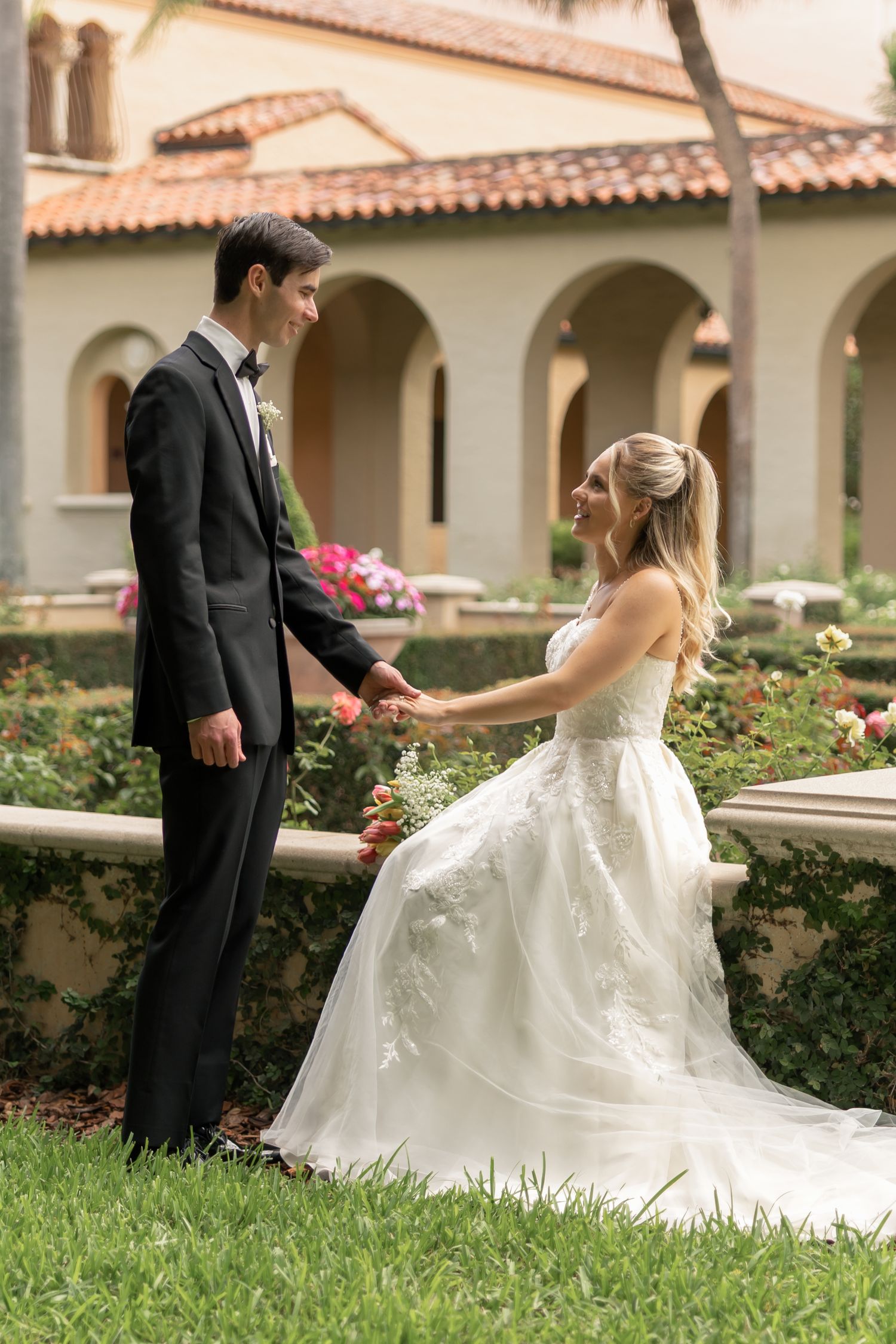 Bride in white lace gown sits on garden wall holding hands with groom in black tuxedo at elegant venue.