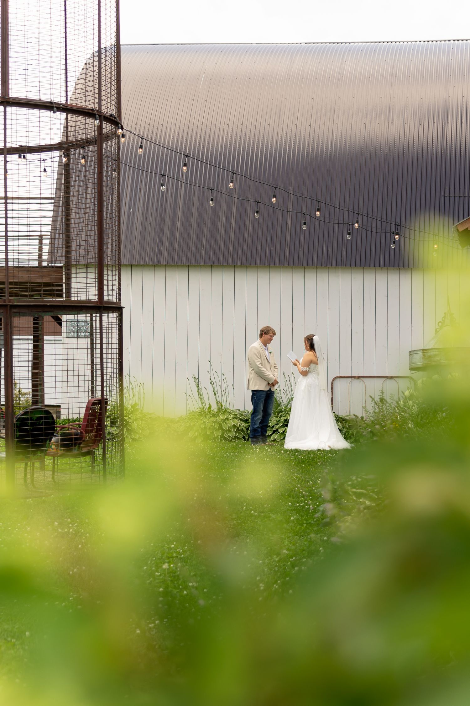 A bride and groom stand together in a lush garden beside a rustic barn adorned with string lights on their wedding day.