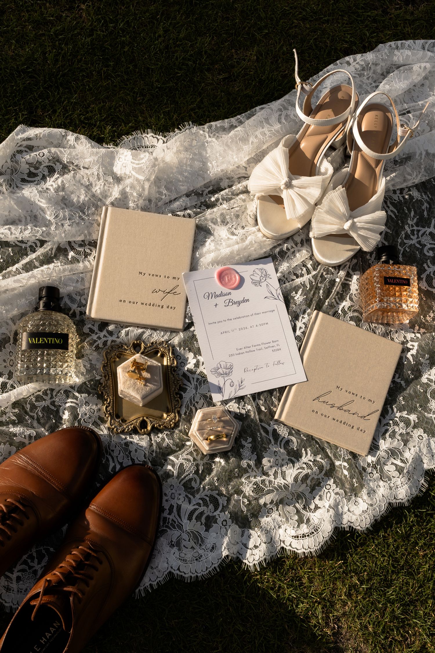 Elegant wedding flat lay with bridal heels, Valentino perfume, invitation, vow books, rings, and brown dress shoes on lace fabric.