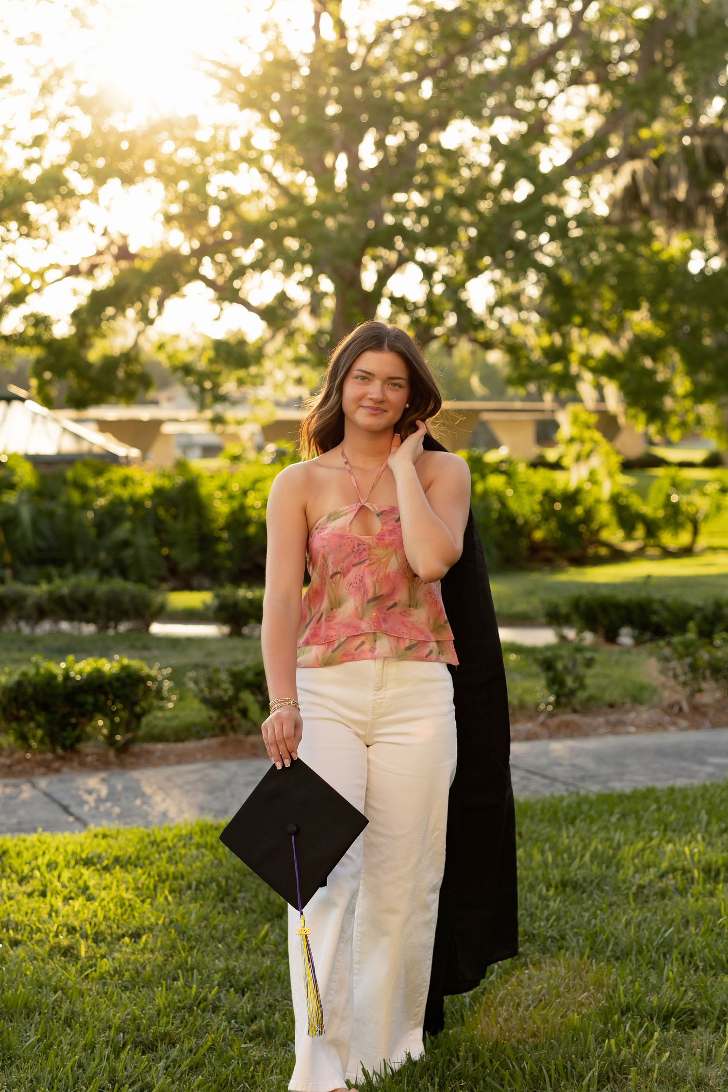 Young woman in floral top and white pants holds graduation cap and gown outdoors in sunny green park setting.