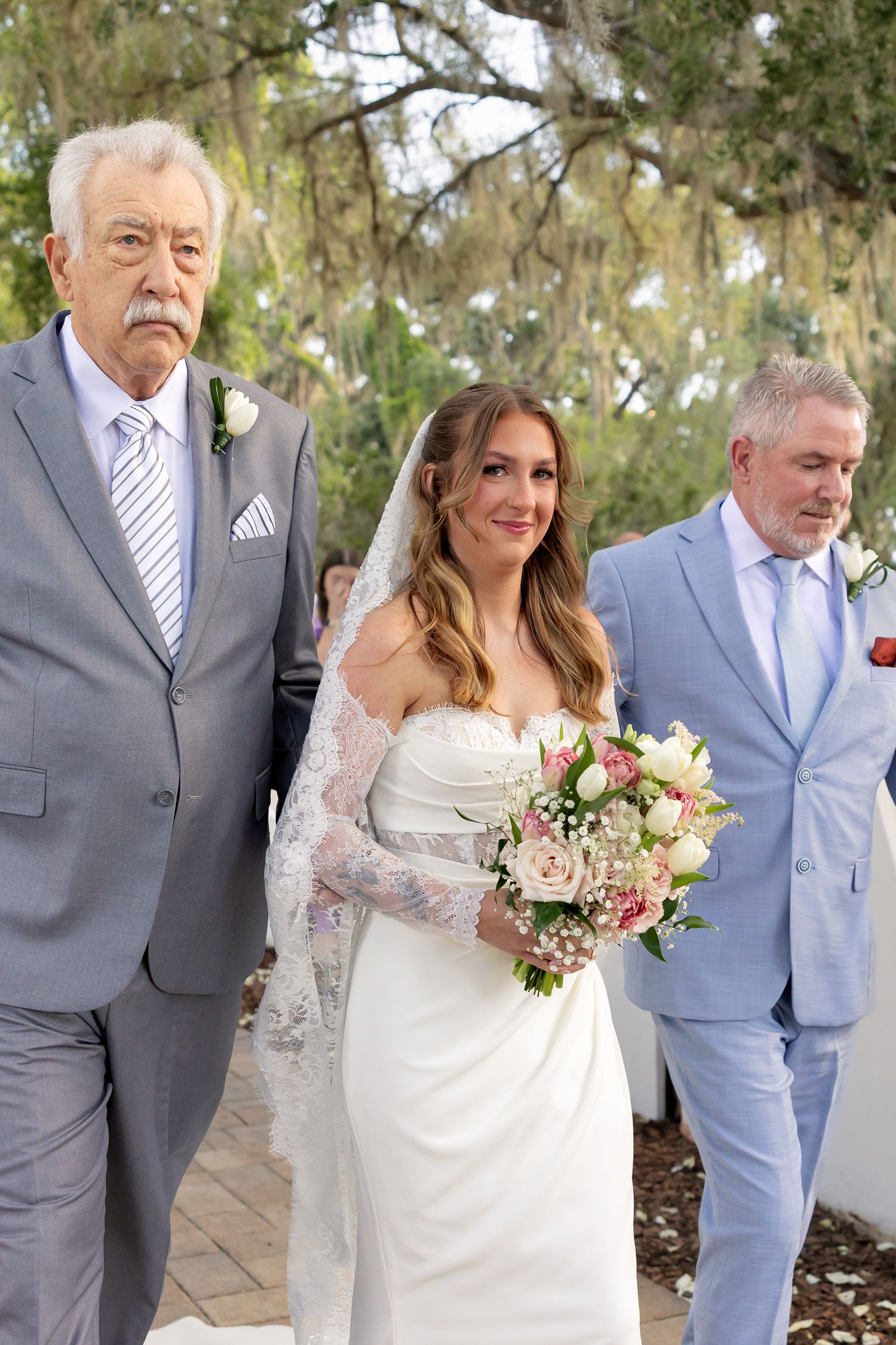Bride in white strapless gown and lace veil holds pink bouquet while being escorted by two men in gray suits.