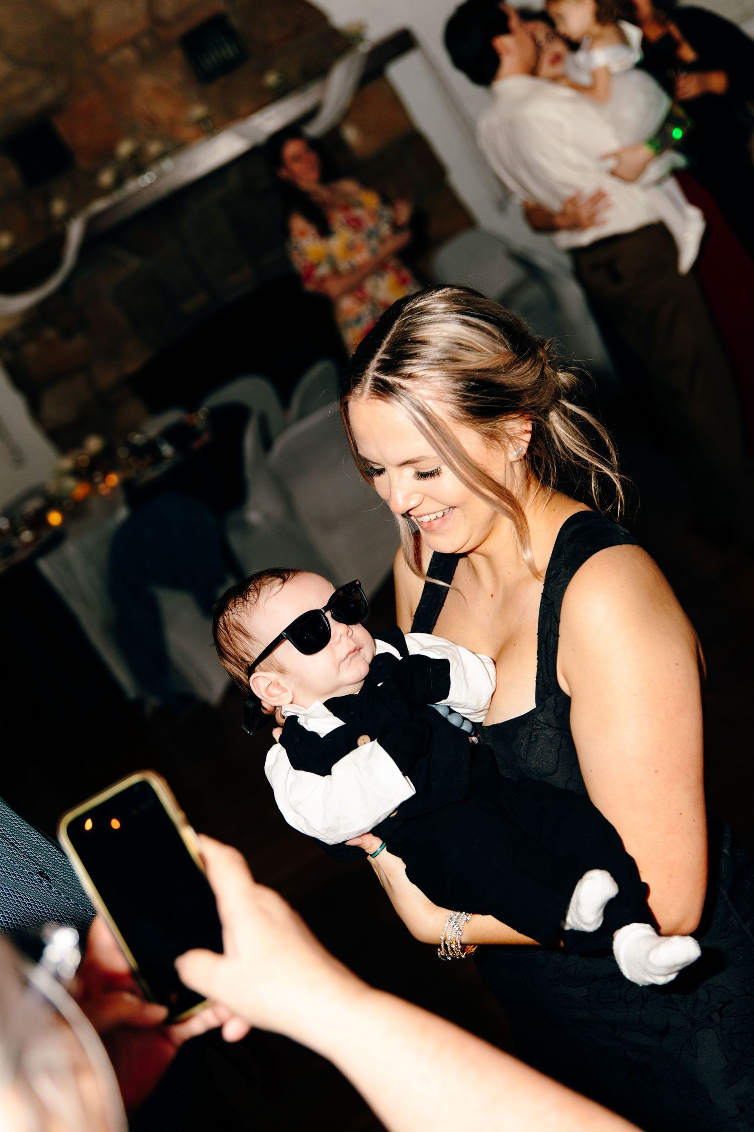 A smiling woman in a black dress holds a tiny baby dressed in a tuxedo and sunglasses at a wedding reception.