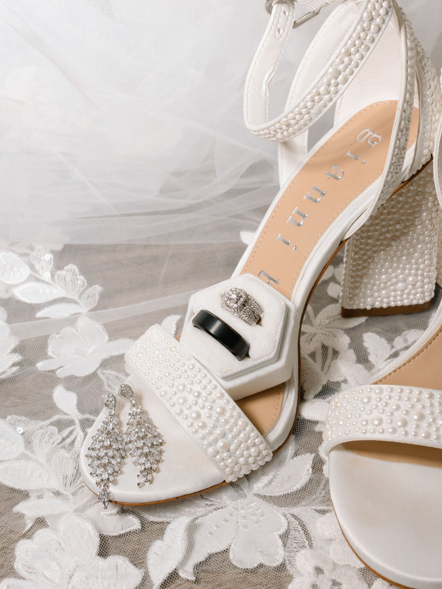 Elegant bridal shoes decorated with pearls and rhinestones displayed alongside delicate white lace fabric.