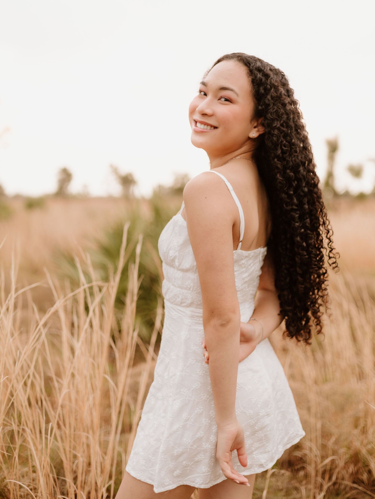 A person in a white sundress laughs joyfully while standing in a golden field of tall grass at sunset.
