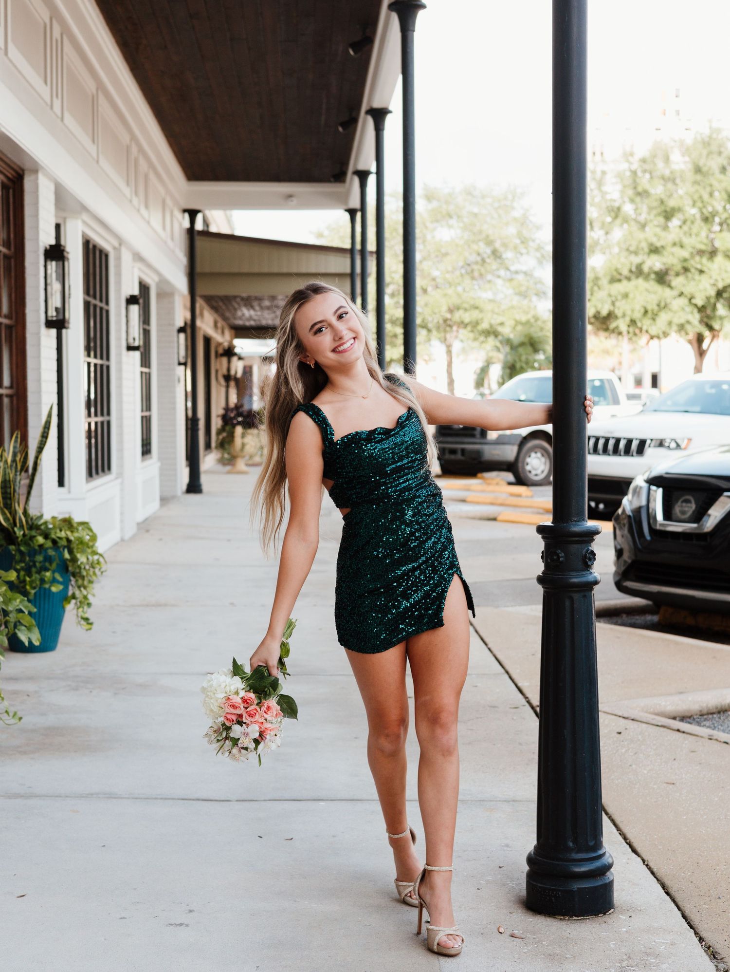 A person in a sparkly green dress poses playfully against a white building column with covered walkway.