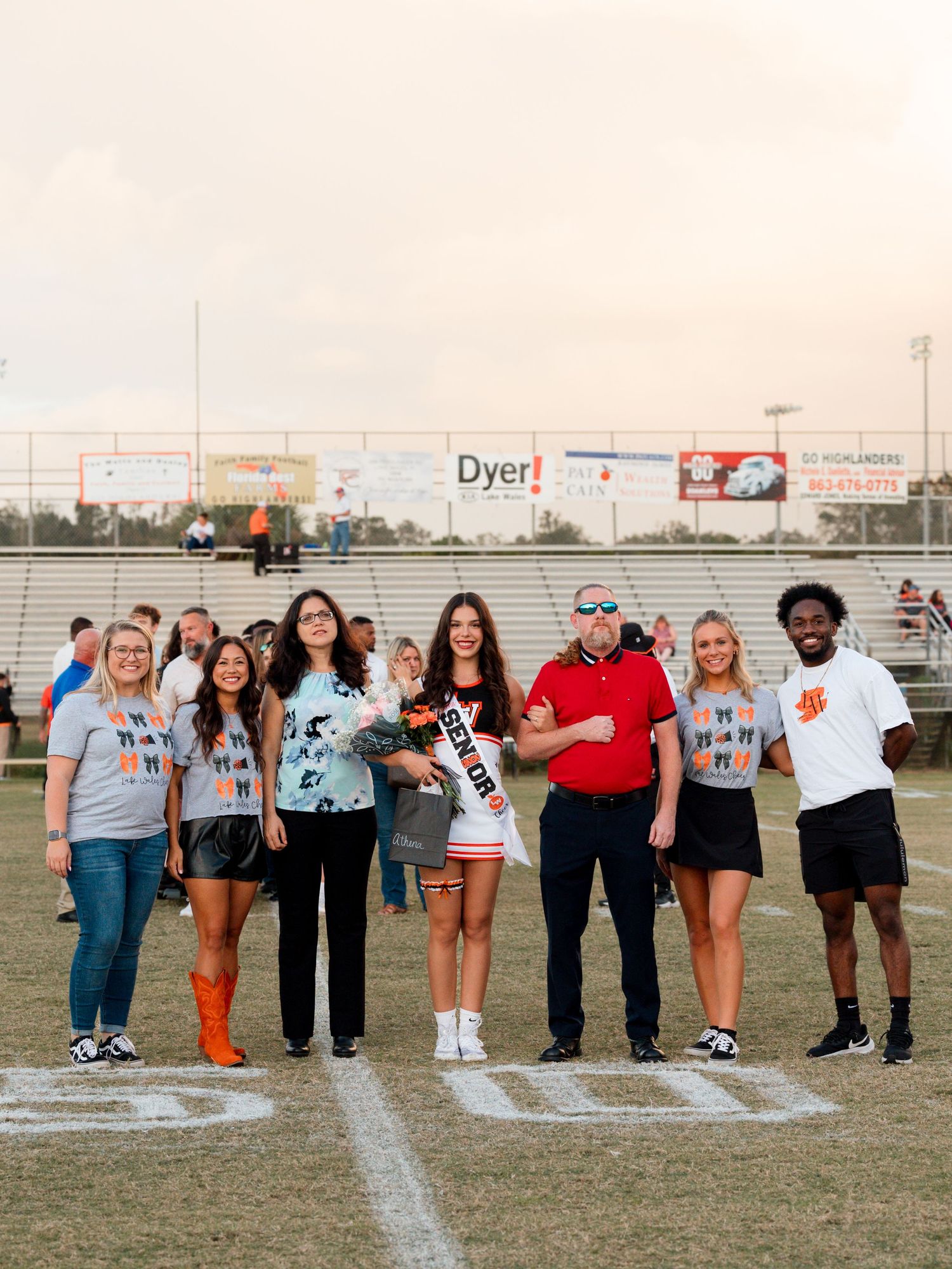 A group of cheerleaders and athletes line up on a sports field in matching team uniforms and casual wear.