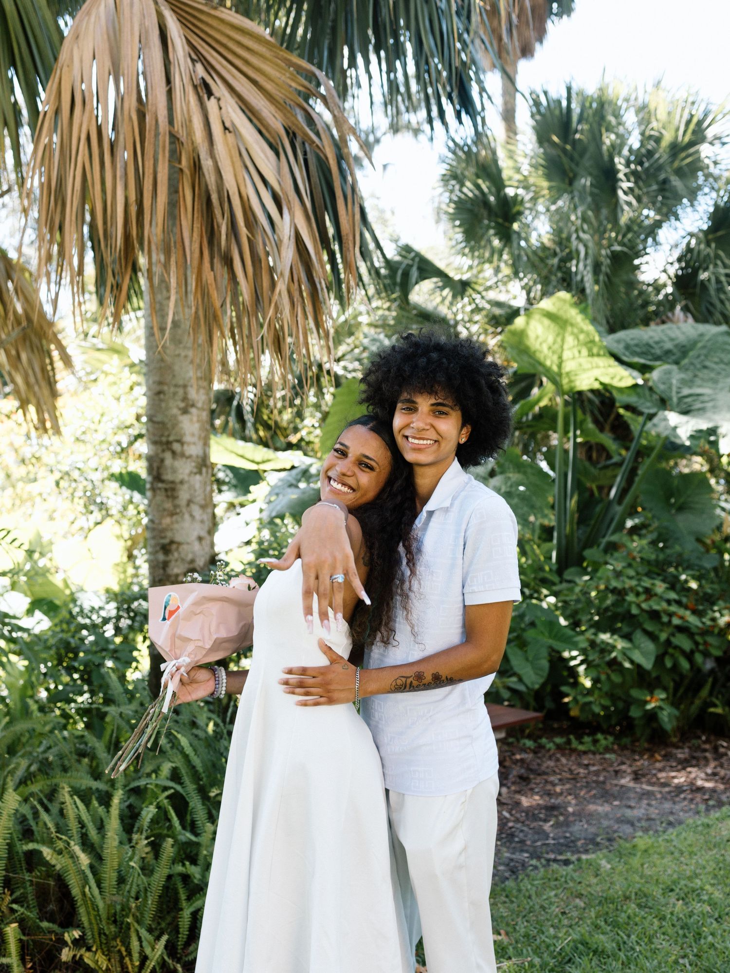 Two people in white outfits embrace and smile while standing in a tropical garden setting with palm trees.