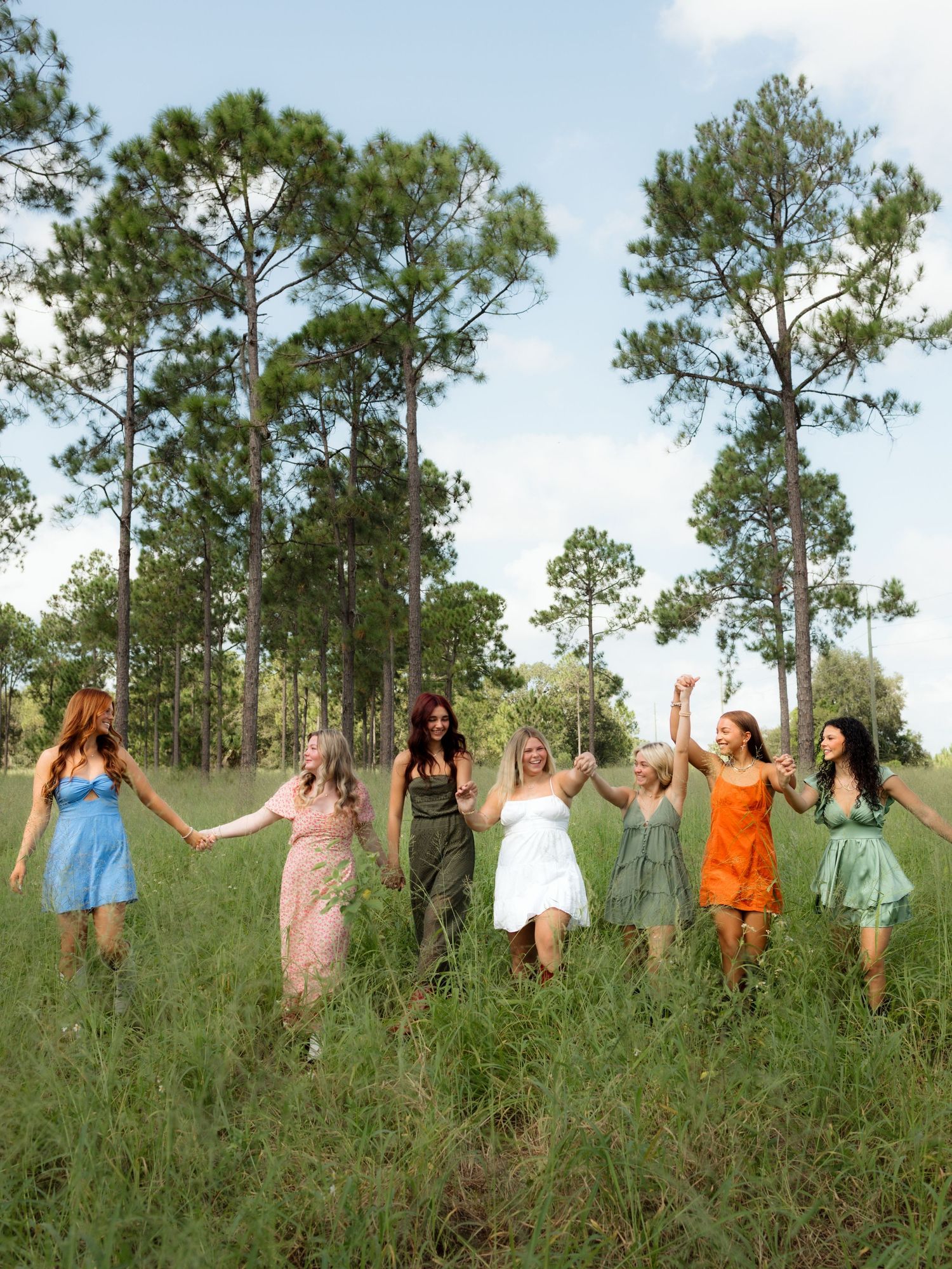 A group in colorful summer dresses hold hands while walking through a field with tall pine trees in the background.